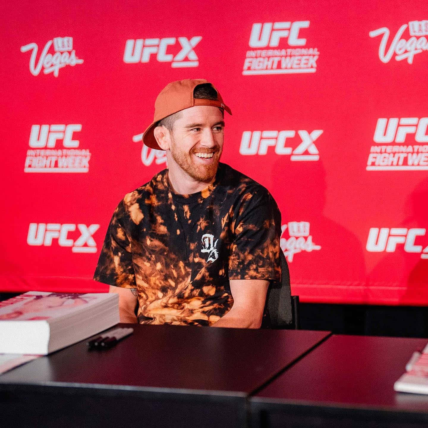MMA 桌布 壁紙 — 一位微笑的拳手穿著紮染襯衫和橙色帽子，在紅色品牌背景的UFC活動中簽名。