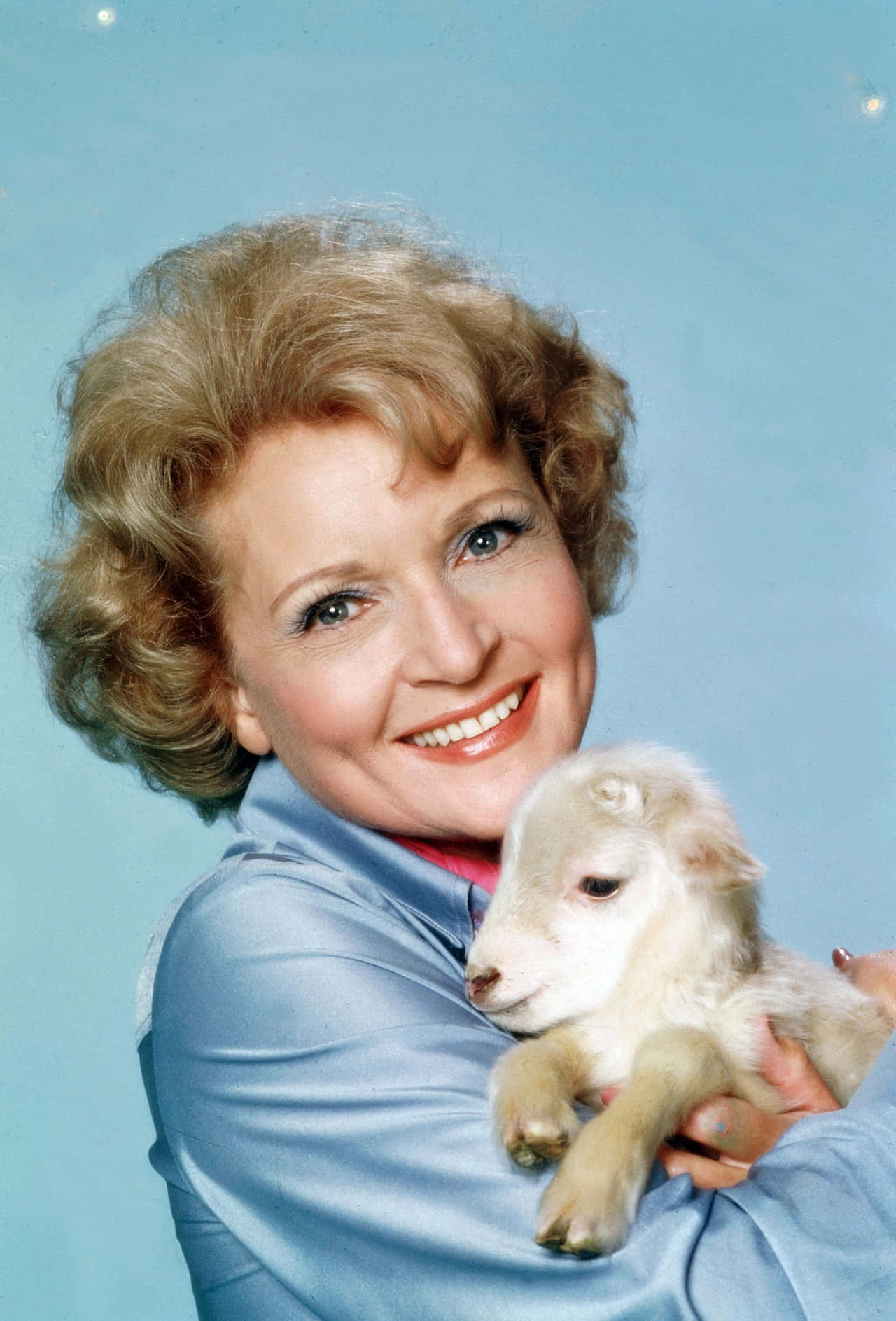 Betty White 桌布 壁紙 — 一位微笑的捲曲金髮女性在柔和的藍色工作室背景前抱著一隻小白羊。