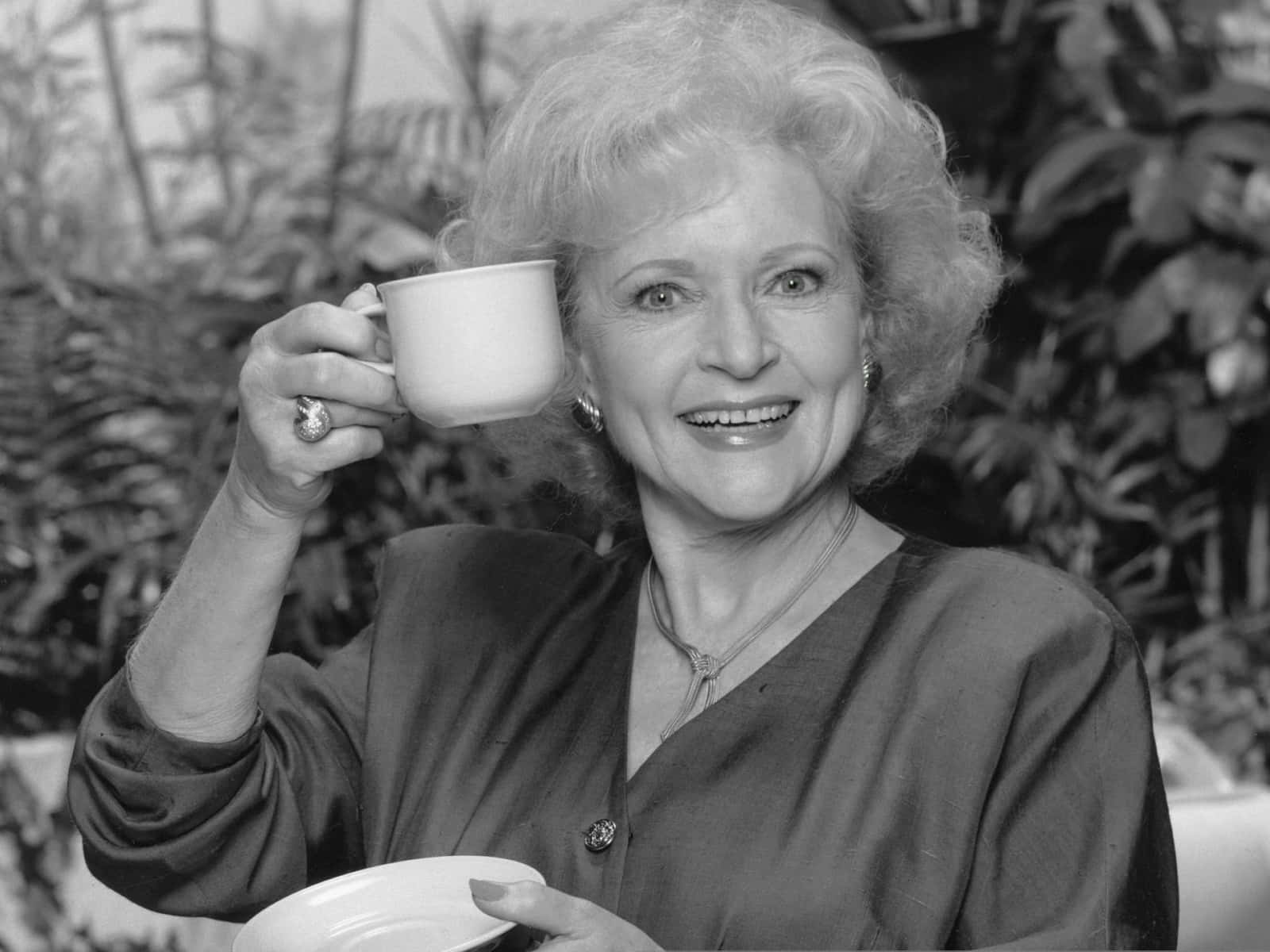 Betty White 桌布 壁紙 — 一位微笑的捲髮年長女性，坐在茂密的樹葉中，手持茶杯和碟子，黑白肖像。
