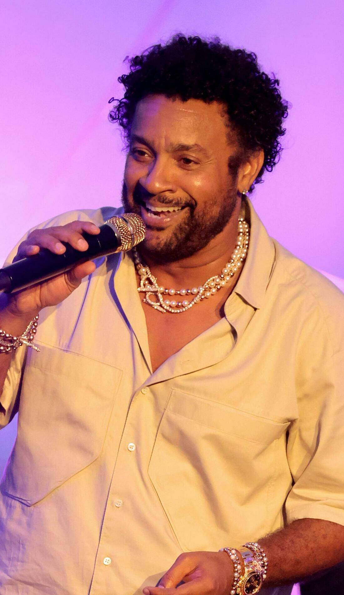 Celebrity 桌布 壁紙 — 一位微笑的捲髮男子在紫色舞台燈光下，戴著多層珍珠項鍊和手鐲對著麥克風唱歌。