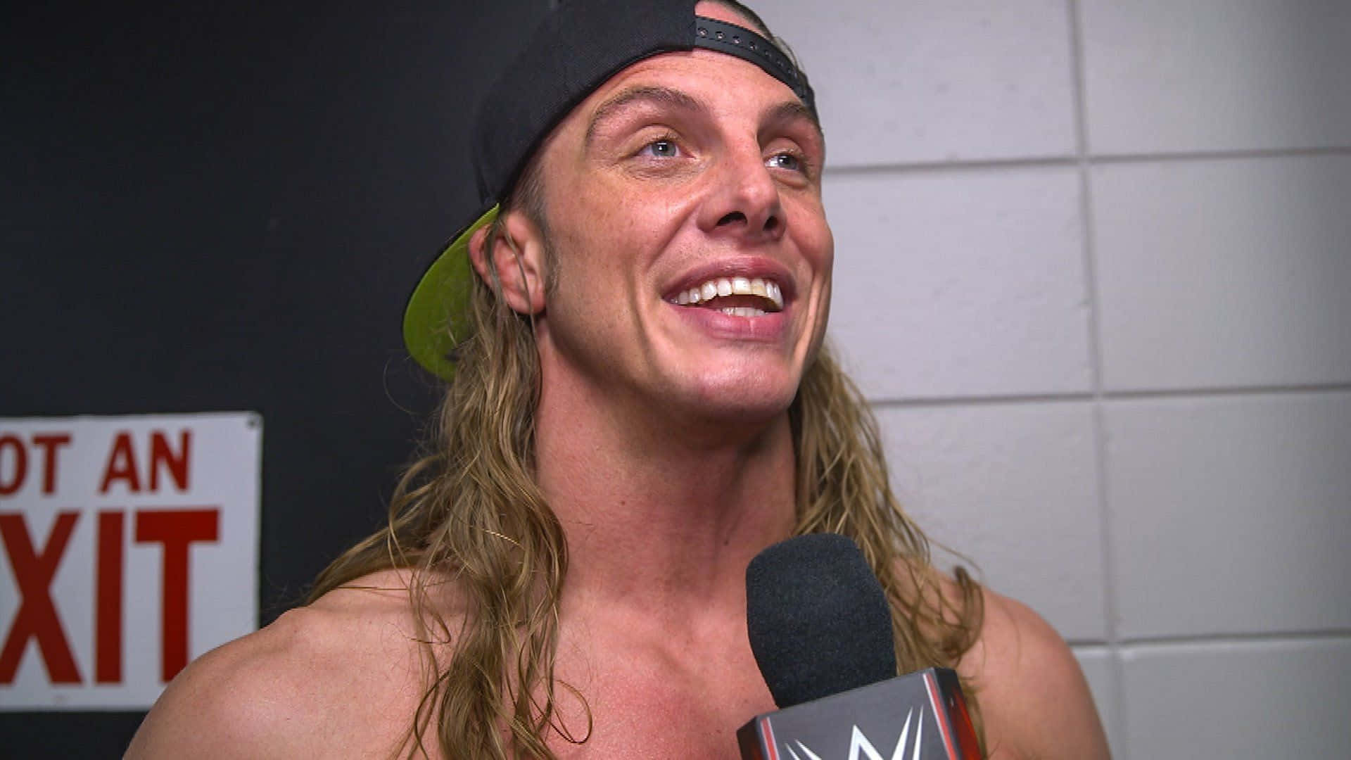 WWE 桌布 壁紙 — 一位微笑的摔角手，長長的金髮和黑色帽子，對著WWE麥克風說話，背景是瓷磚牆和紅色出口標誌。