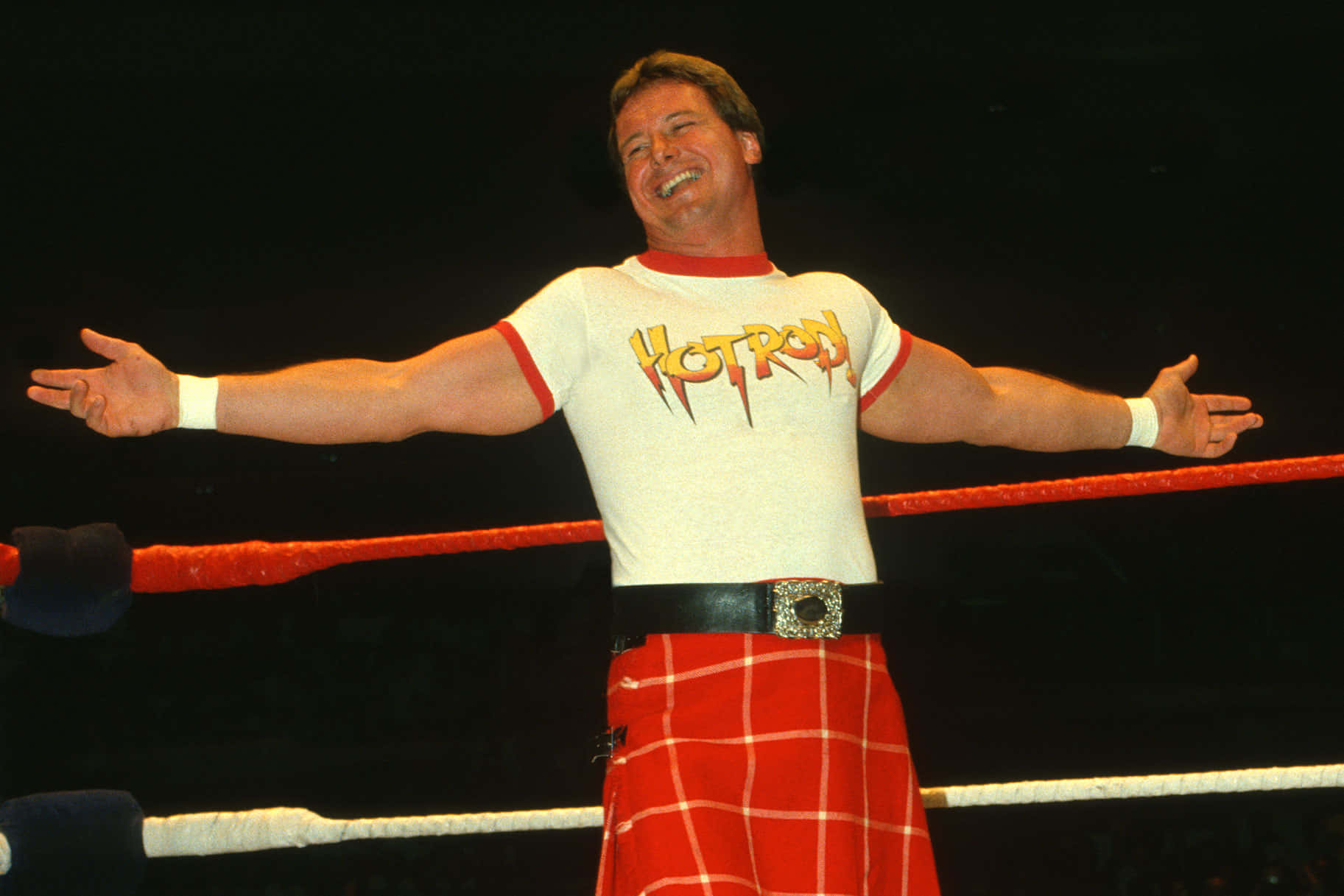 WWE 桌布 壁紙 — 一位微笑的摔角手穿著紅色格子裙和「Hotrod」襯衫，在明亮的燈光下於摔角場中張開雙臂站立。