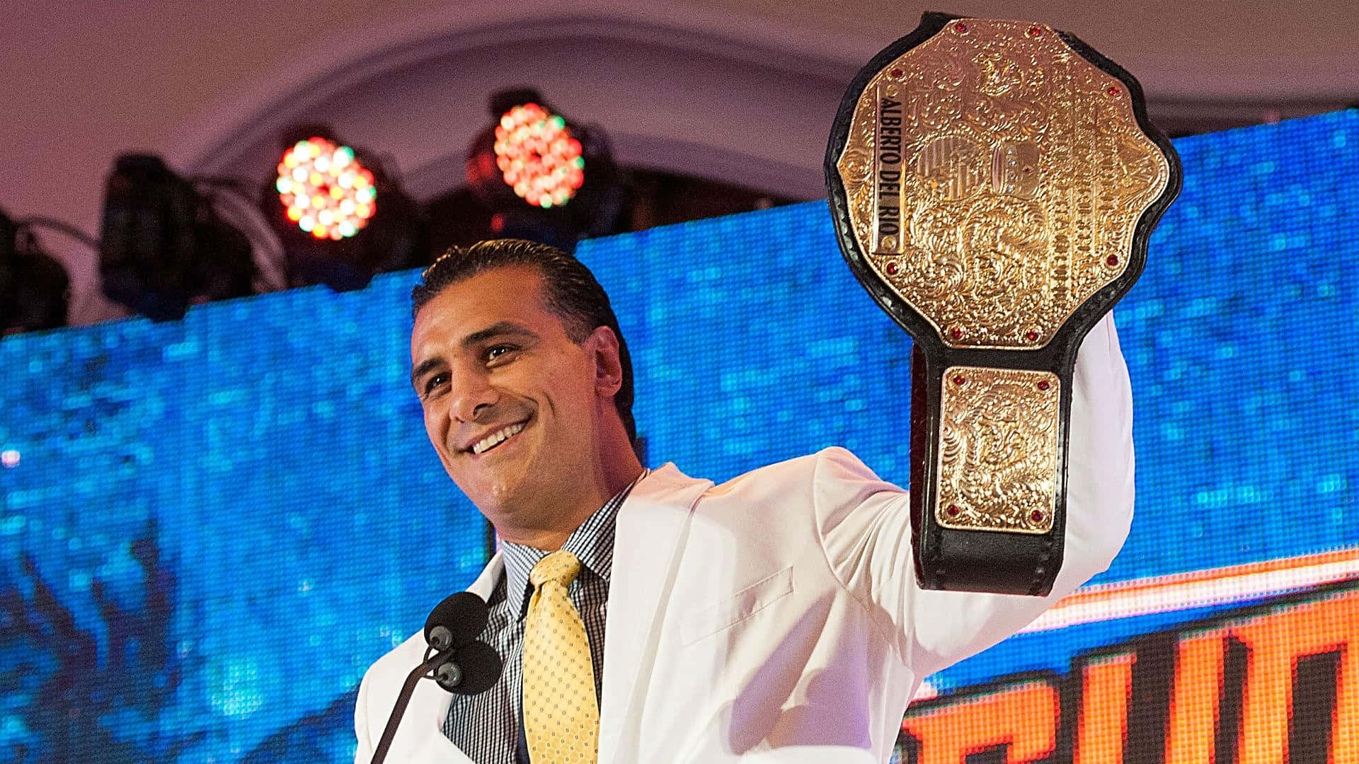 WWE 桌布 壁紙 — 一位微笑的摔角手身穿白色西裝，在充滿活力的藍色LED舞台背景下自豪地舉起一條金色冠軍腰帶。