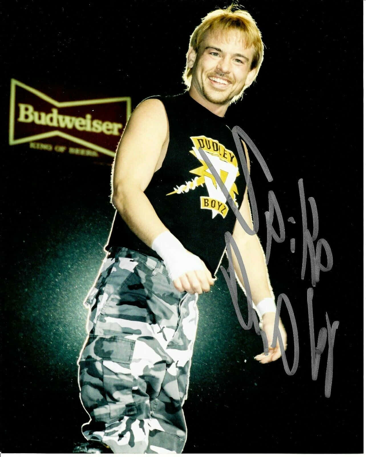 Athlete 桌布 壁紙 — 一位微笑的摔角手身穿黑色背心和迷彩褲，在帶有Budweiser標誌的深色背景前自信地擺姿勢。