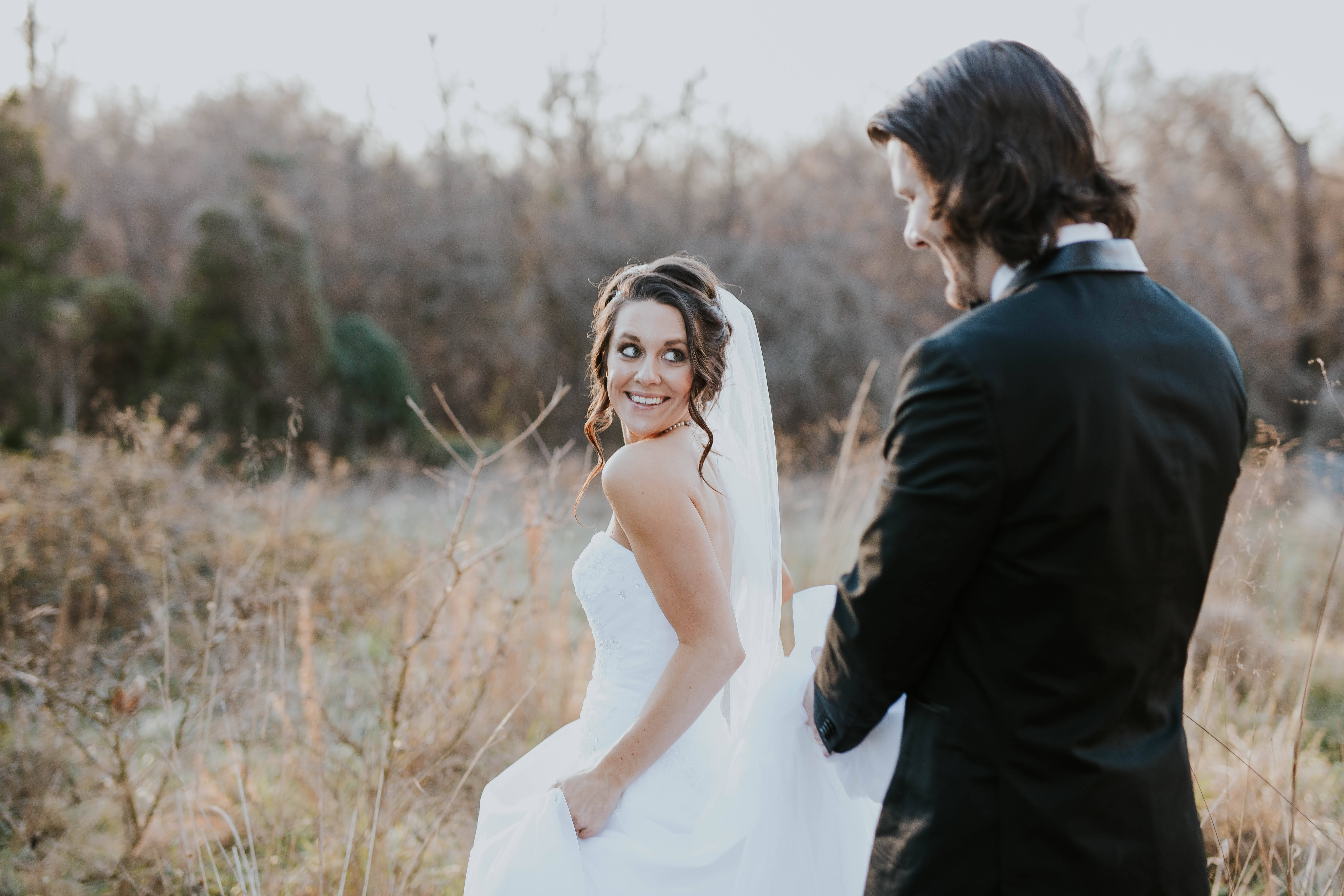 Groom 桌布 壁紙 — 一位微笑的新娘穿著白色禮服，轉身凝視著站在陽光普照的乾草地上的黑色西裝新郎。