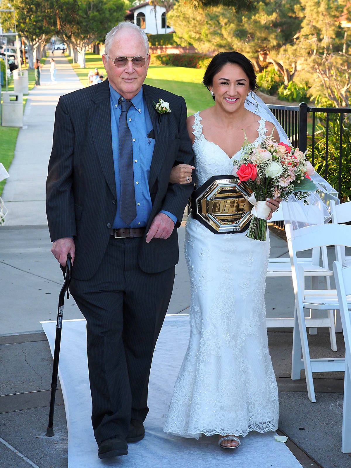 Champion 桌布 壁紙 — 一位微笑的新娘穿著白色蕾絲婚紗，與手持拐杖的父親在明亮的日光下沿著戶外走道行走，兩人均穿著正式服裝。