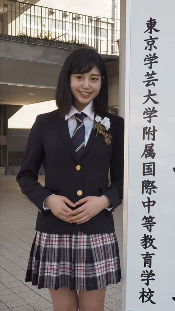 美學 桌布 壁紙 — 一位微笑的日本女學生，身穿深色西裝外套和格子裙，站在一個有漢字標誌的標牌旁邊。