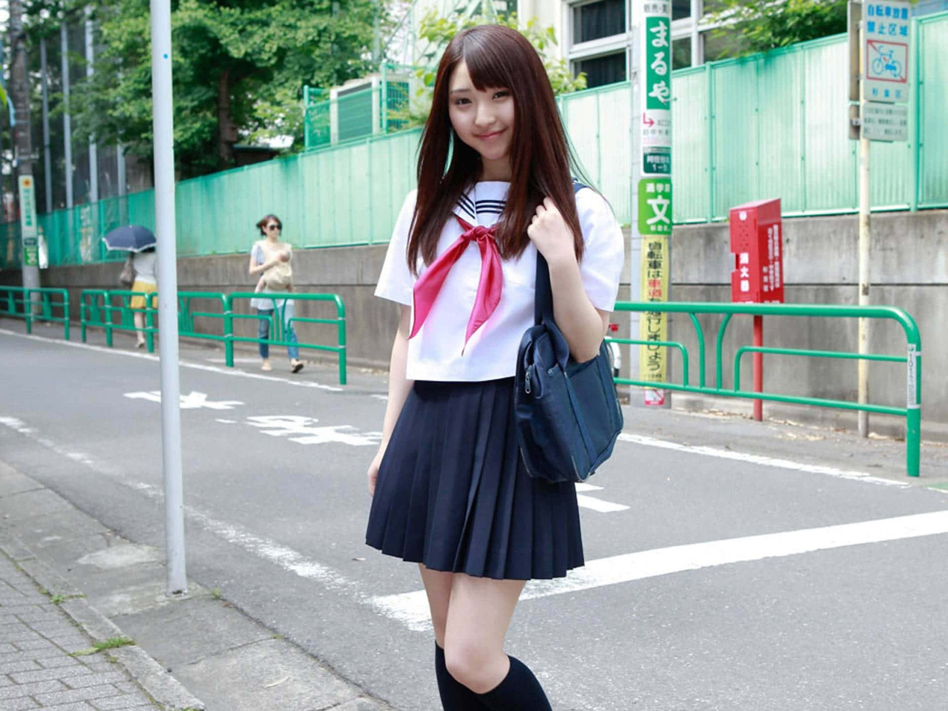 Japanese School Girl 桌布 壁紙 — 一位微笑的日本女學生，身穿白色水手服，搭配紅色蝴蝶結和海軍藍百褶裙，站在城市人行道上，手持一個海軍藍手提袋。