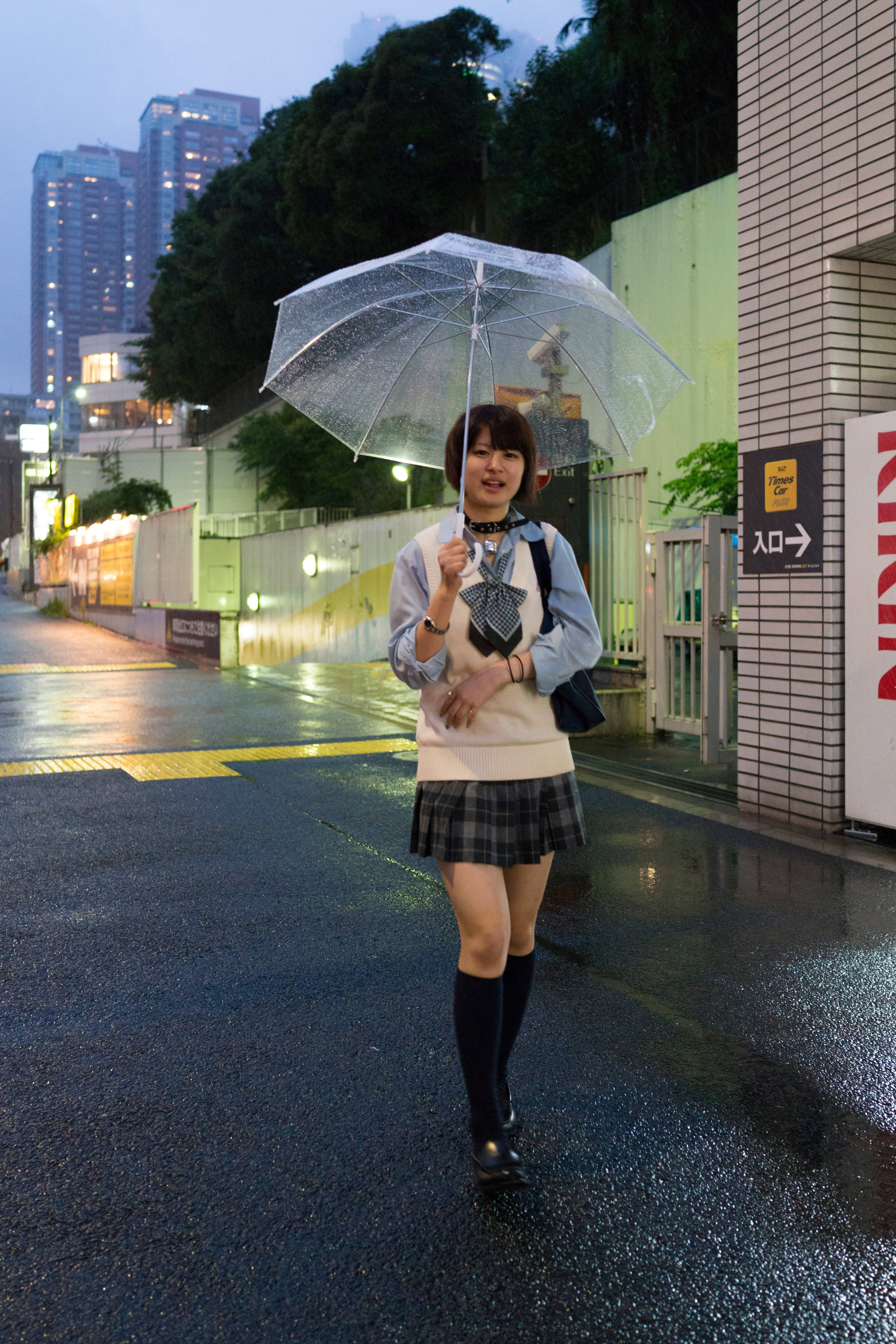 Raining 桌布 壁紙 — 一位微笑的日本女學生穿著制服，在黃昏時分撐著透明雨傘走在被雨水打濕的城市街道上。