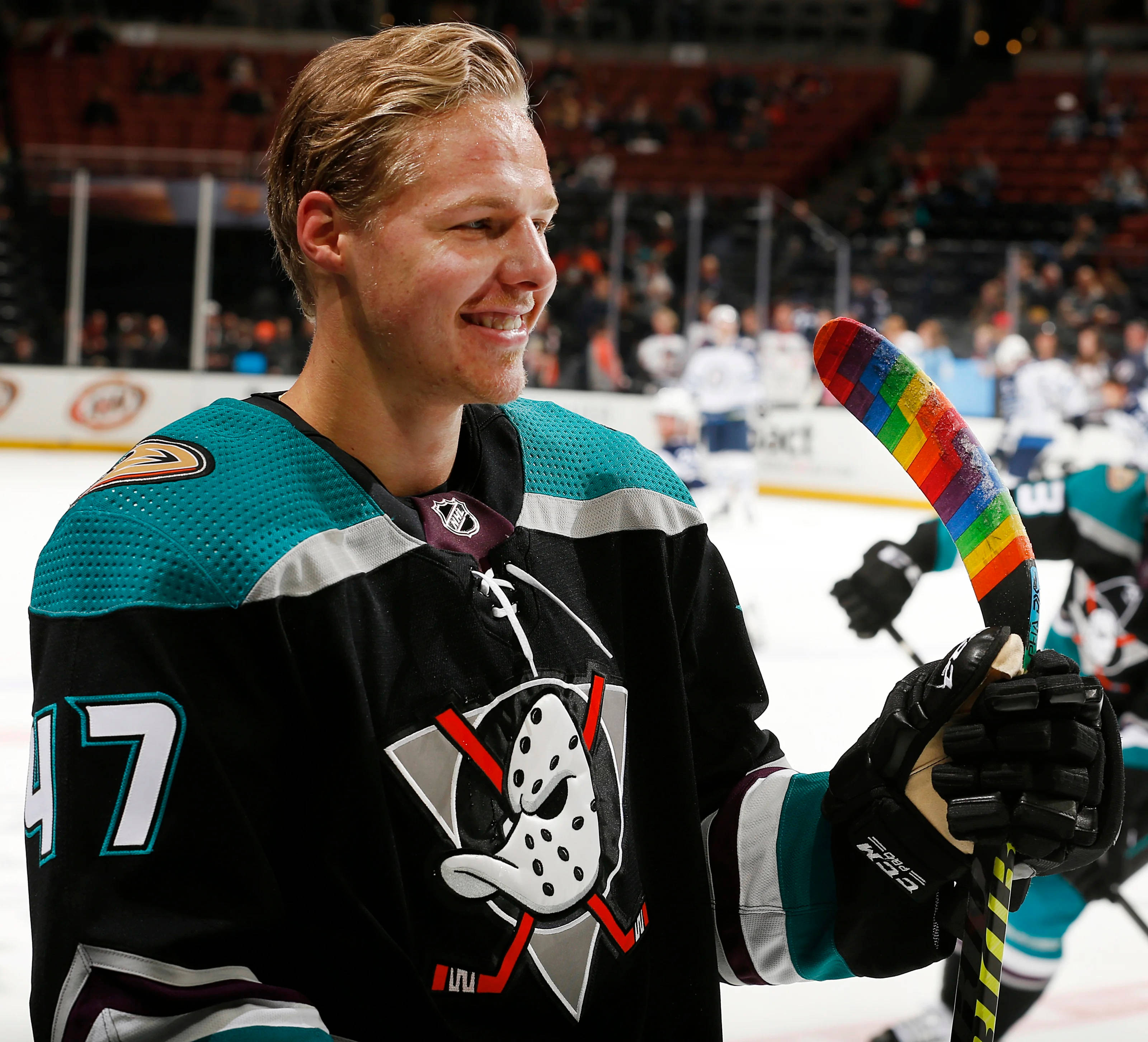 hockey 桌布 壁紙 — 一位微笑的曲棍球運動員穿著青綠色和黑色球衣，在冰場上拿著彩虹色的球棍，背景有觀眾。
