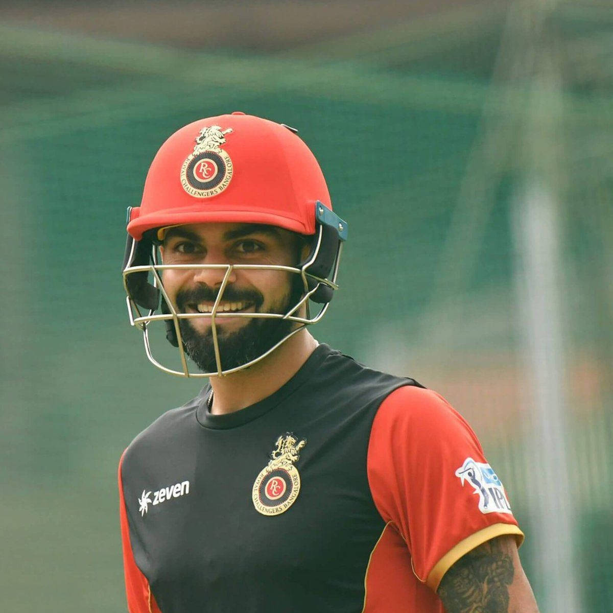 royal challengers bangalore 桌布 壁紙 — 一位微笑的板球運動員，戴著紅色頭盔，身穿黑紅色球衣，自信地站在綠色場地上。
