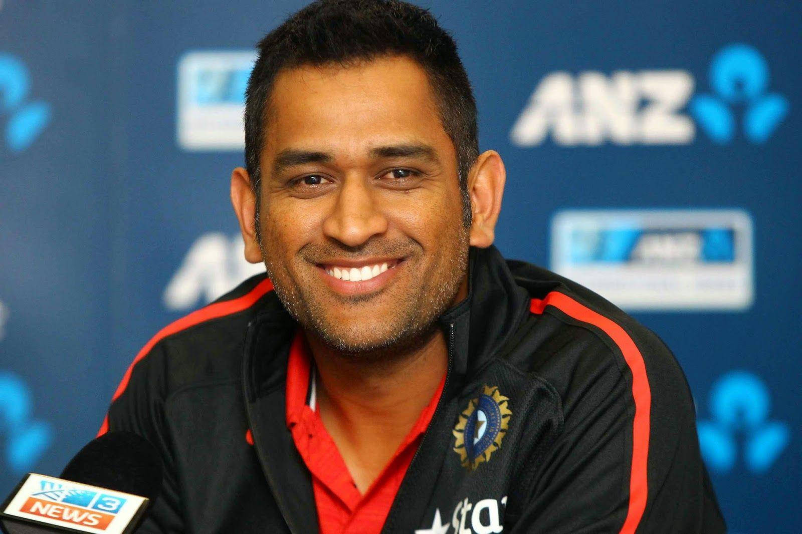 Ms Dhoni 桌布 壁紙 — 一位微笑的板球運動員穿著帶有印度標誌的黑色和紅色運動夾克，在藍色品牌背景的新聞發布會上。