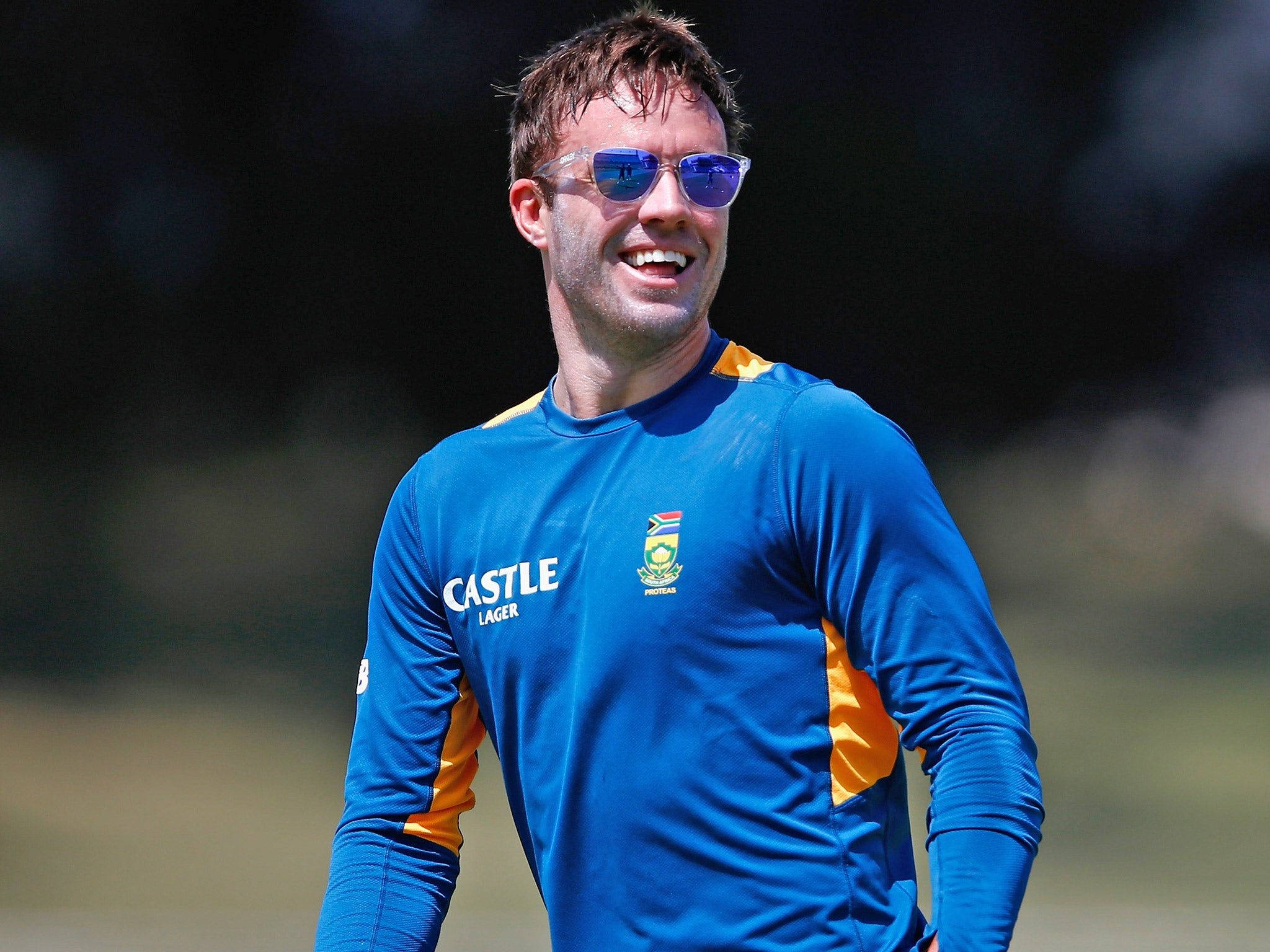 Ab De Villiers 桌布 壁紙 — 一位微笑的板球運動員穿著藍色和橙色的訓練球衣，戴著太陽眼鏡，在陽光明媚的日子裡站在戶外。