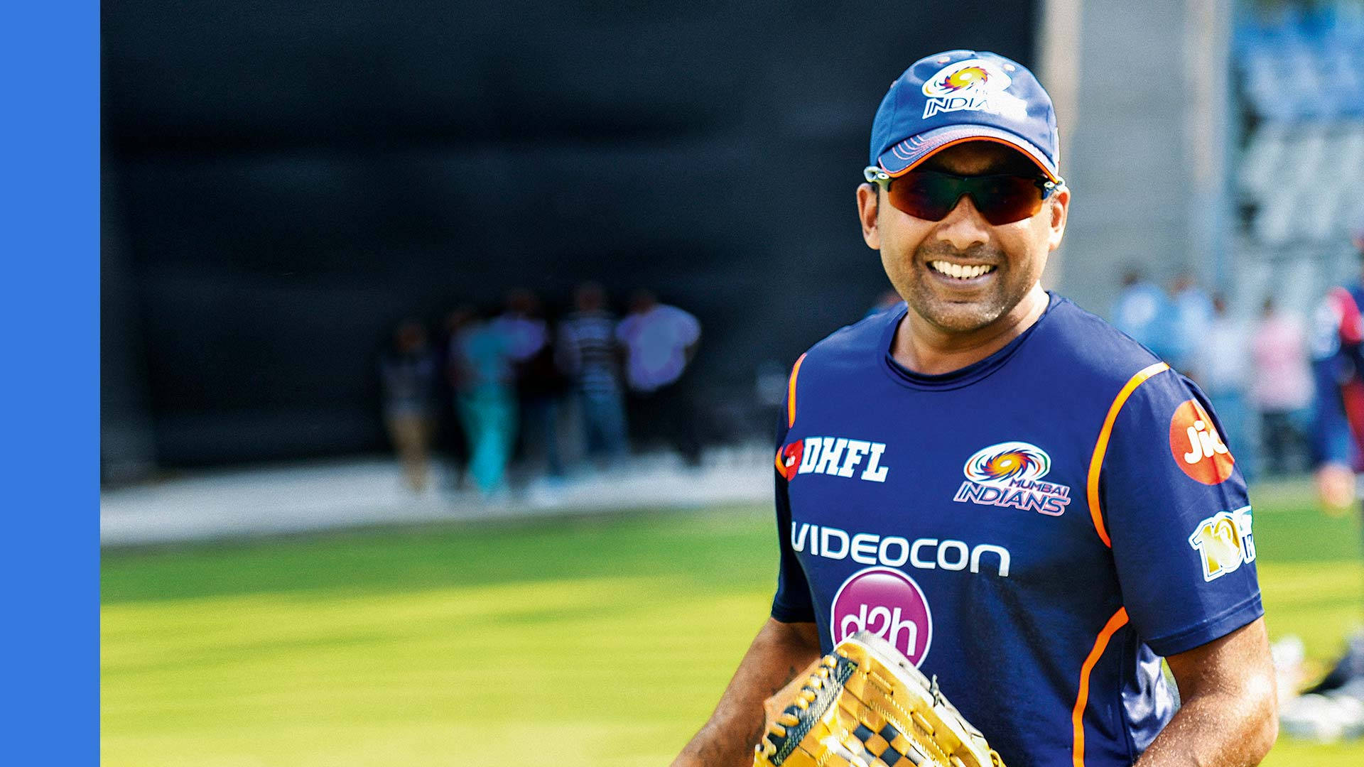 Sri Lanka Cricket 桌布 壁紙 — 一位微笑的板球運動員身穿藍色Mumbai Indians球衣和帽子，在陽光明媚的球場上拿著黃色手套。