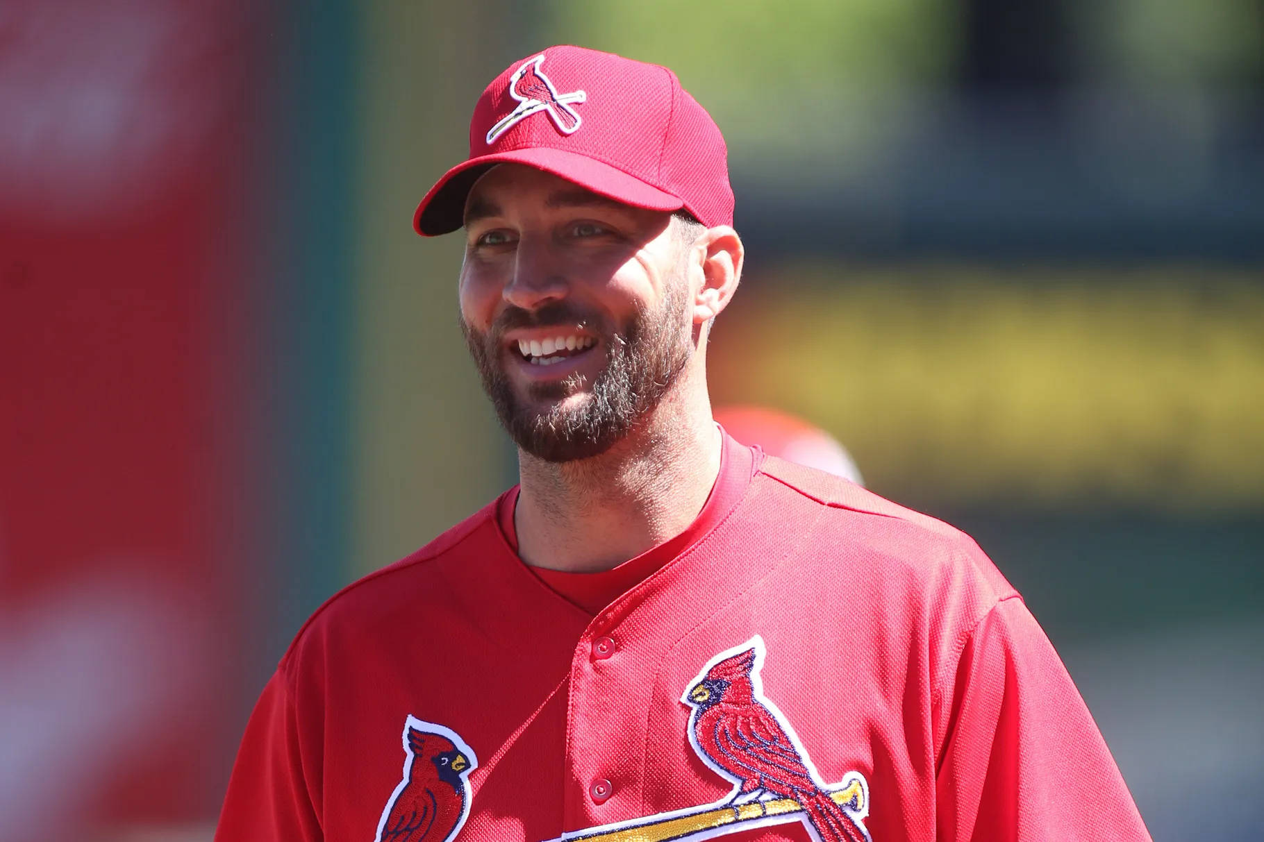 Adam Wainwright 桌布 壁紙 — 一位微笑的棒球運動員，身穿紅色 Cardinals 球衣和帽子，在明亮的陽光下站在戶外。