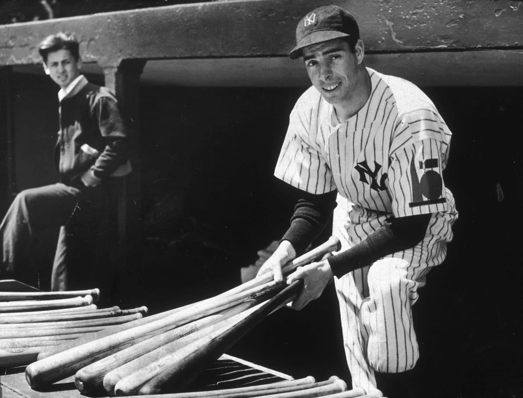 Joe Dimaggio 桌布 壁紙 — 一位微笑的棒球運動員身穿條紋制服，在球員休息區拿著球棒，身後坐著另一名男子。