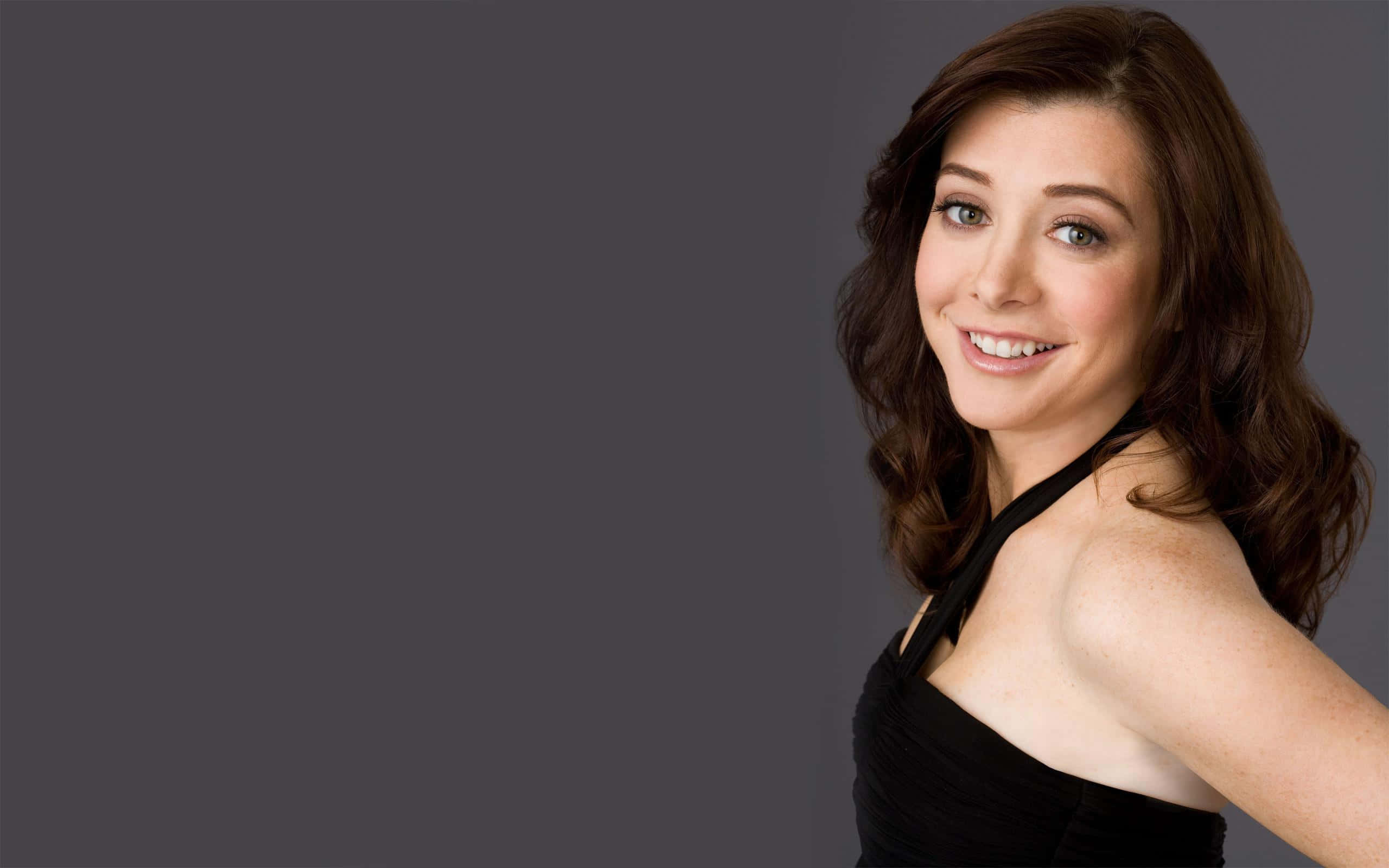 Alyson Hannigan 桌布 壁紙 — 一位微笑的棕色頭髮、綠色眼睛的女性，穿著黑色掛頸上衣，背景是純灰色。