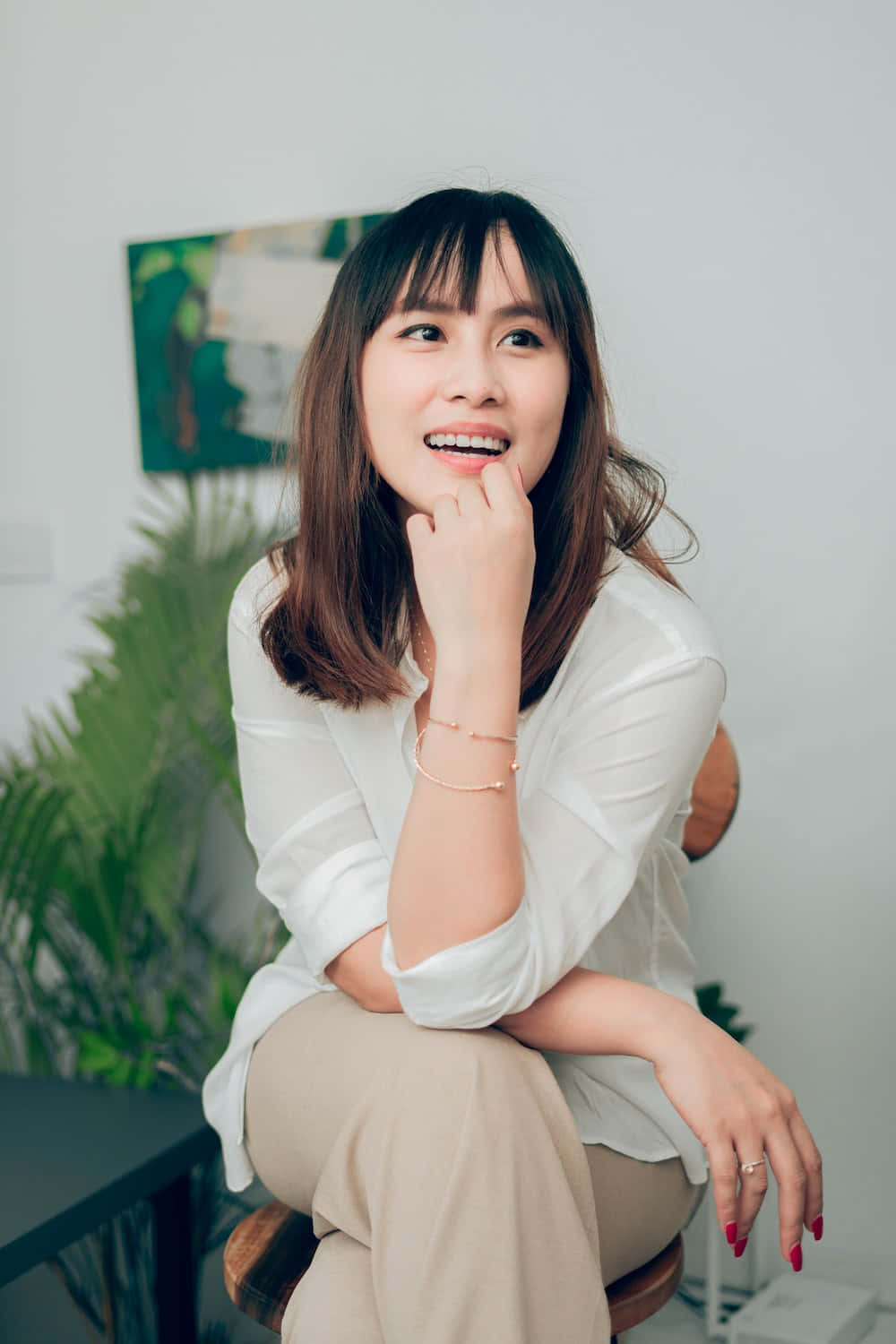 美女 桌布 壁紙 — 一位微笑的棕髮女性坐在明亮的室內空間的凳子上，身後有綠色植物和現代畫作，若有所思。