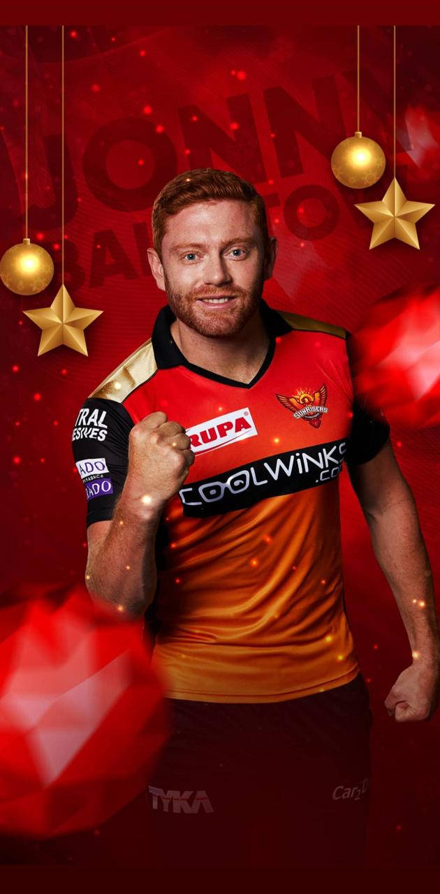 Sunrisers Hyderabad 桌布 壁紙 — 一位微笑的橄欖球聯賽球員身穿紅金色球衣，在掛有金色裝飾的節日紅色背景前自信地擺姿勢。