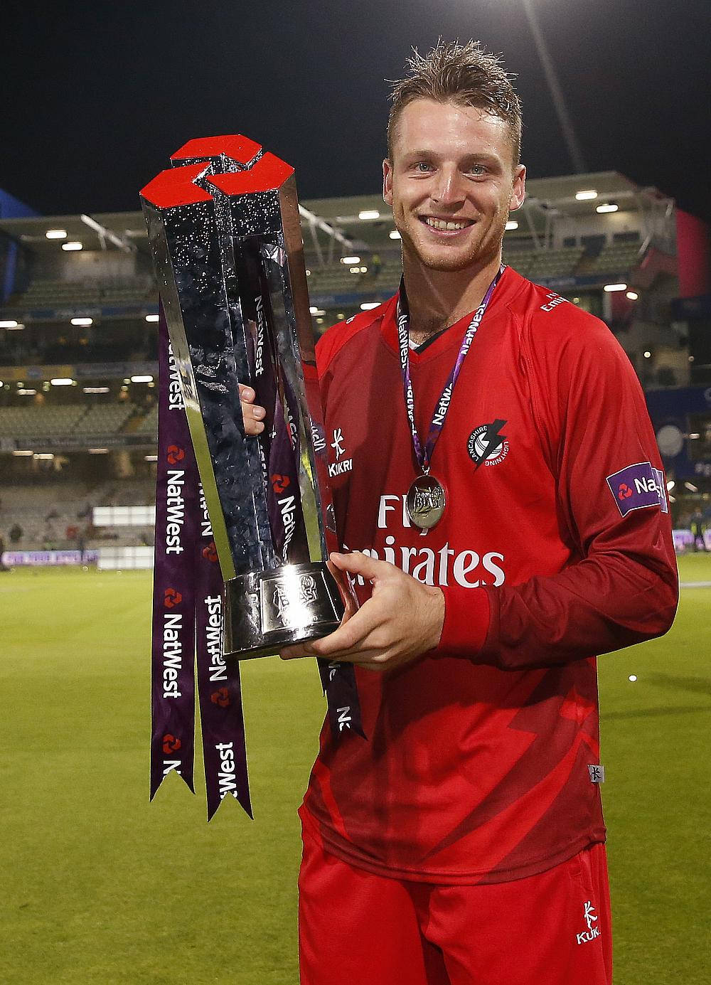 jos buttler 桌布 壁紙 — 一位微笑的橄欖球運動員身穿紅色球衣，在夜晚的體育場上手持一個帶有紫色絲帶的大銀色獎盃。
