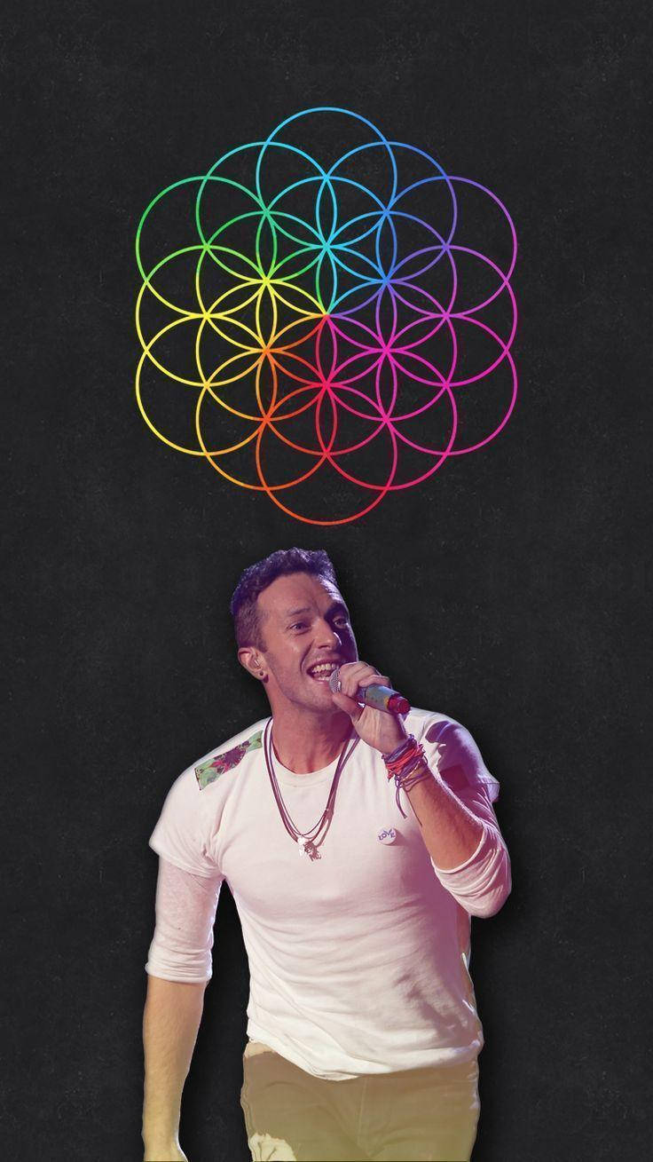 Chris Martin 桌布 壁紙 — 一位微笑的歌手在黑暗背景下，於發光的彩虹生命之花符號下方握著麥克風。