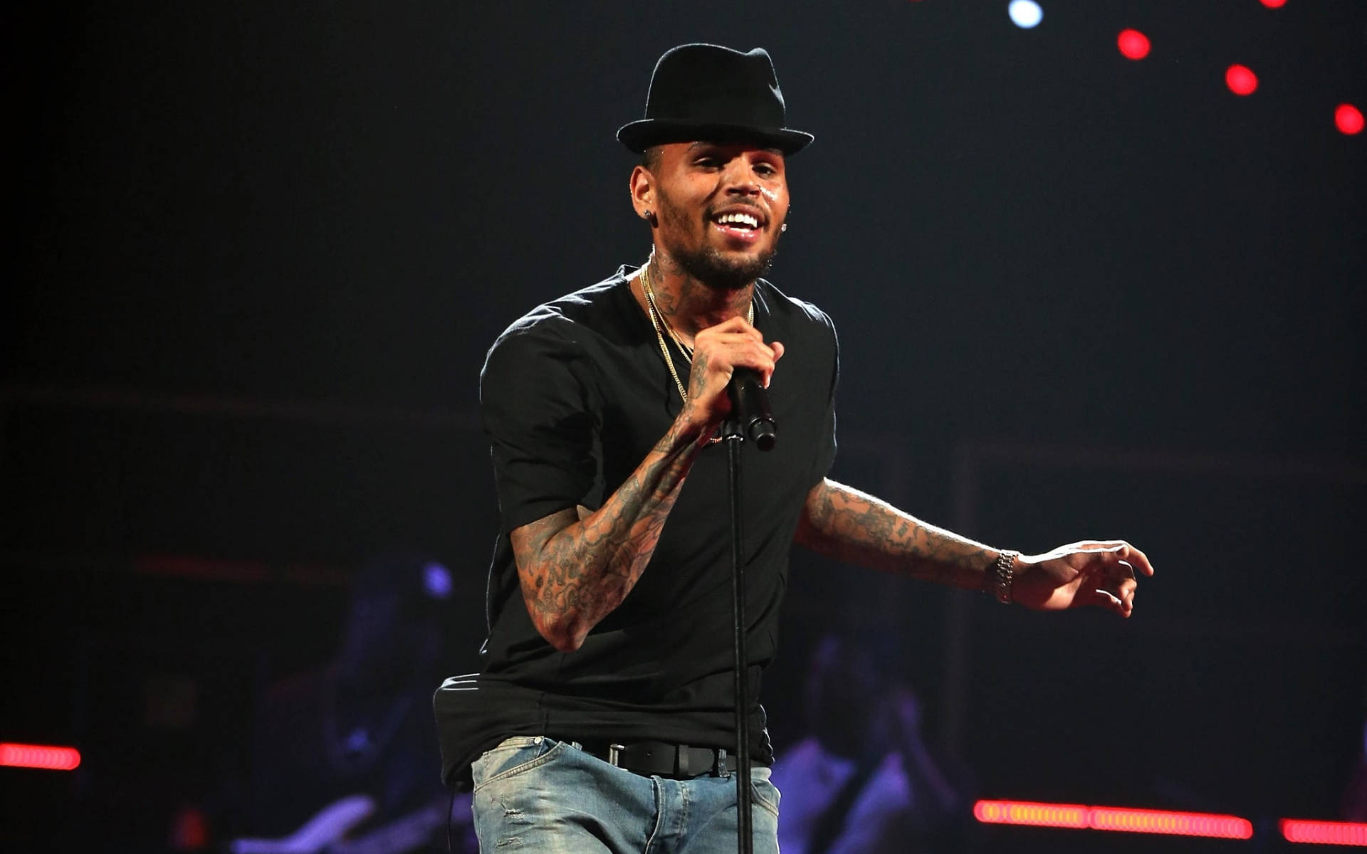 Chris Brown 桌布 壁紙 — 一位微笑的歌手戴著黑色軟呢帽和刺青，在昏暗的舞台上伴隨紅燈充滿活力地表演。