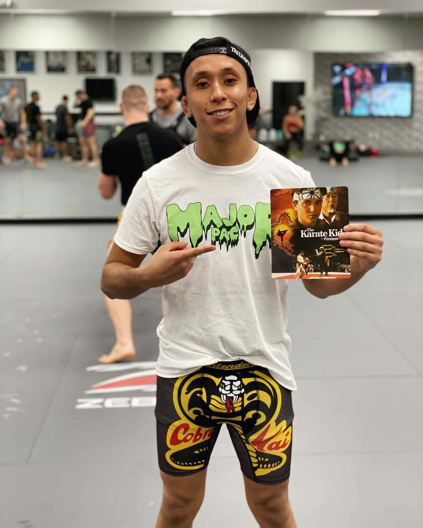 健身房 桌布 壁紙 — 一位微笑的武術家在健身房自豪地舉著《Karate Kid》電影海報，身穿Cobra Kai短褲。