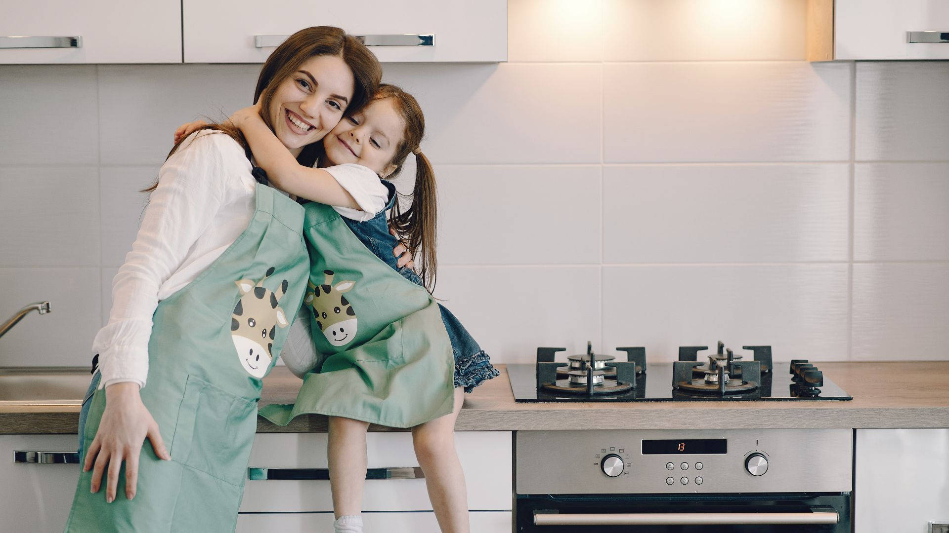 Mom 桌布 壁紙 — 一位微笑的母親和她年幼的女兒在明亮的現代廚房裡穿著相配的綠色長頸鹿圍裙，溫暖地擁抱在一起。