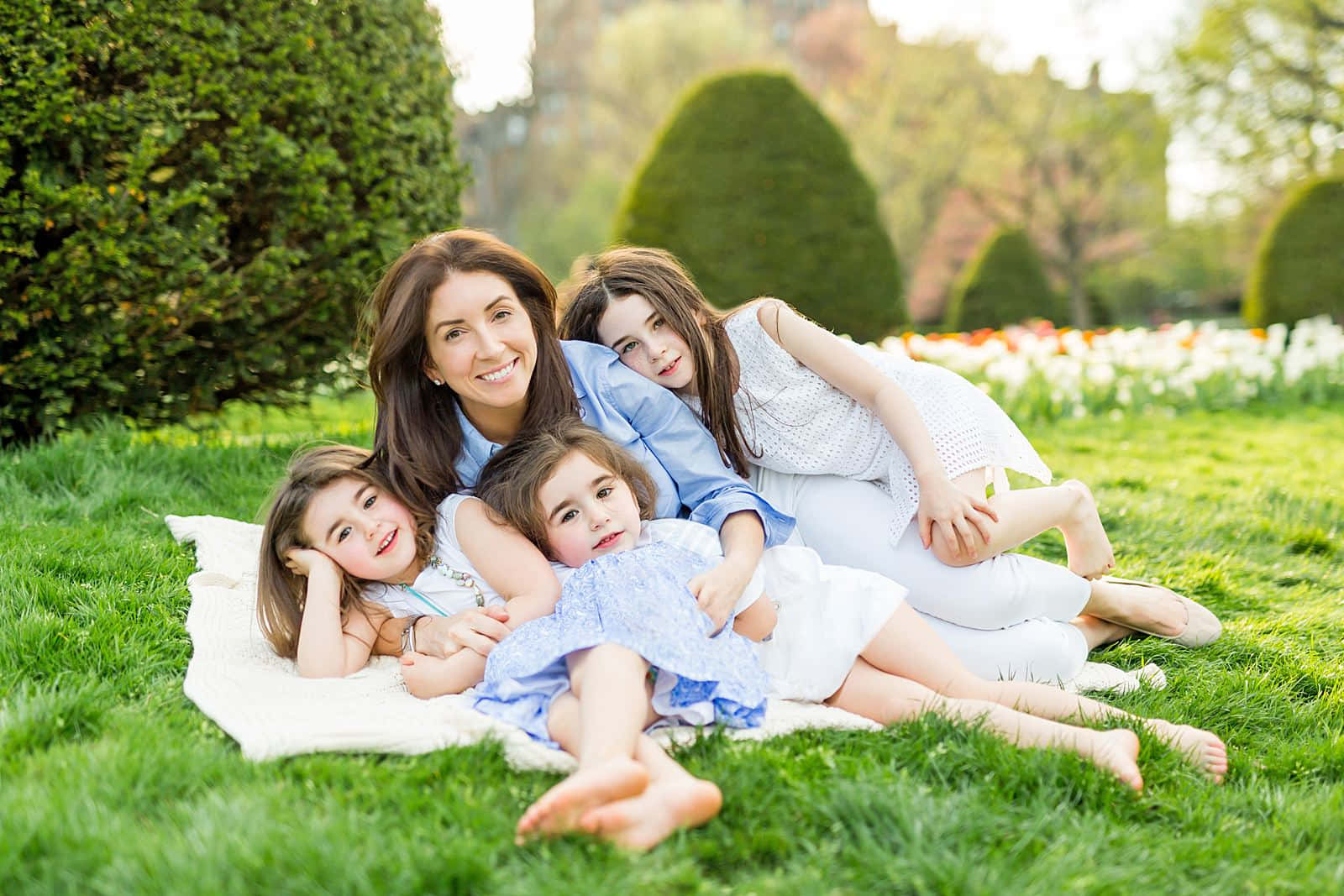 Spring Family 桌布 壁紙 — 一位微笑的母親和她的三個女兒在陽光明媚的公園裡，依偎在白色毯子上，周圍是修剪整齊的樹籬和色彩繽紛的花朵。