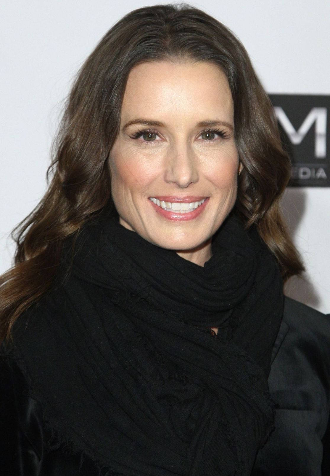 Celebrity 桌布 壁紙 — 一位微笑的波浪棕色頭髮女性，在帶有媒體標誌的白色背景前戴著黑色圍巾。