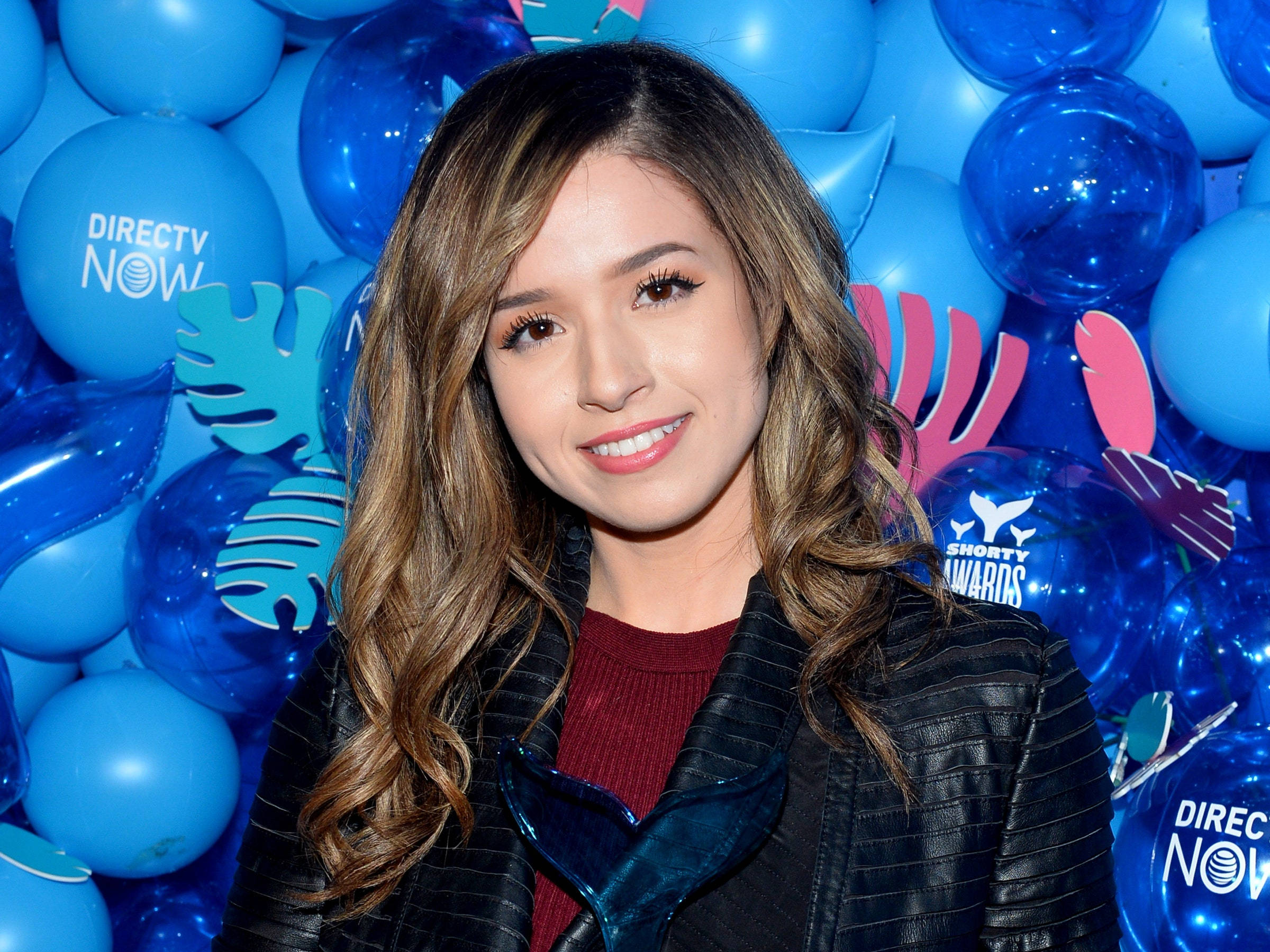 pokimane 桌布 壁紙 — 一位微笑的波浪棕色頭髮女性站在印有DirecTV Now和Shorty Awards標誌的藍色氣球牆前。