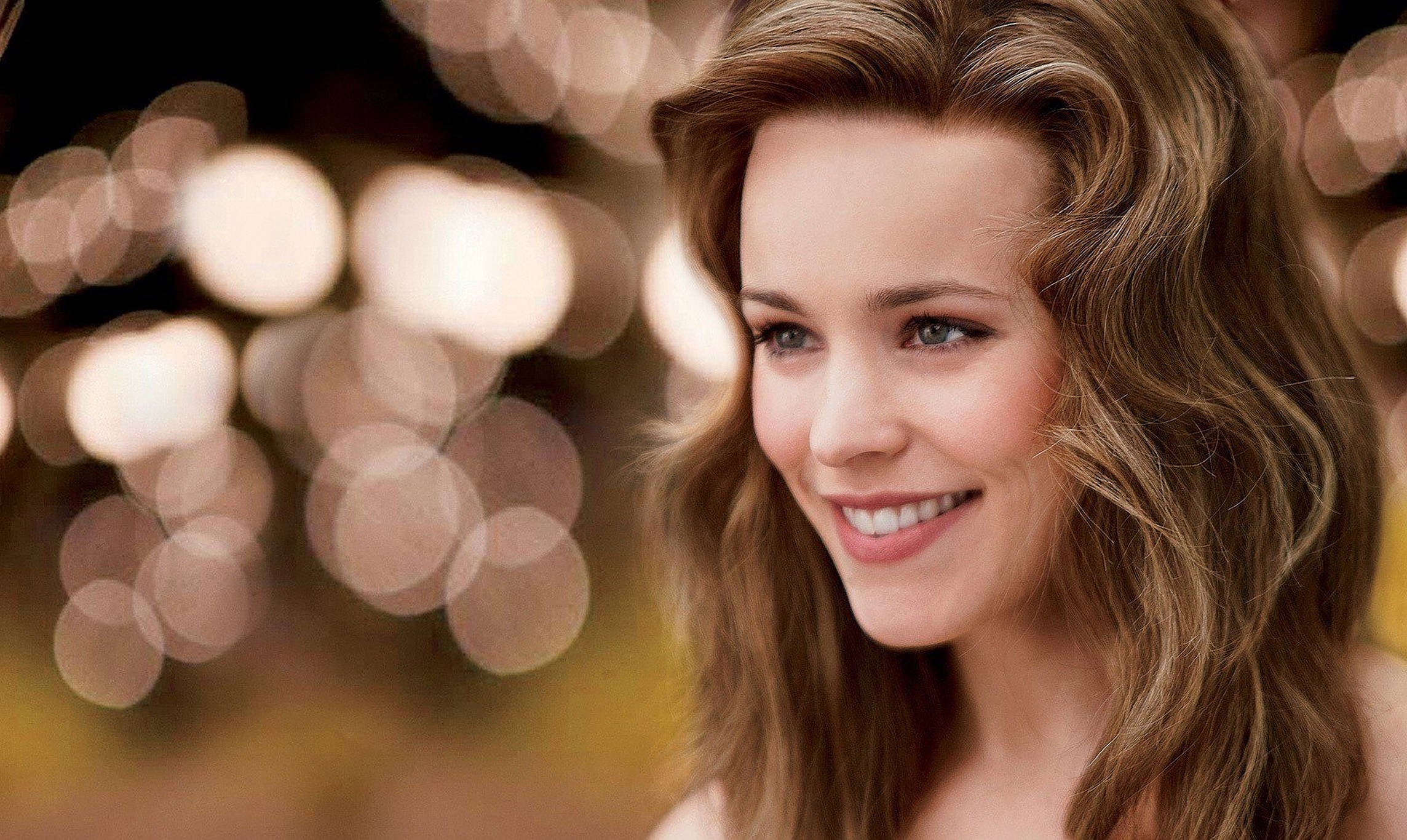 Rachel Mcadams 桌布 壁紙 — 一位微笑的波浪棕色頭髮女性站在柔和的金色光斑前，營造出溫暖發光的背景。