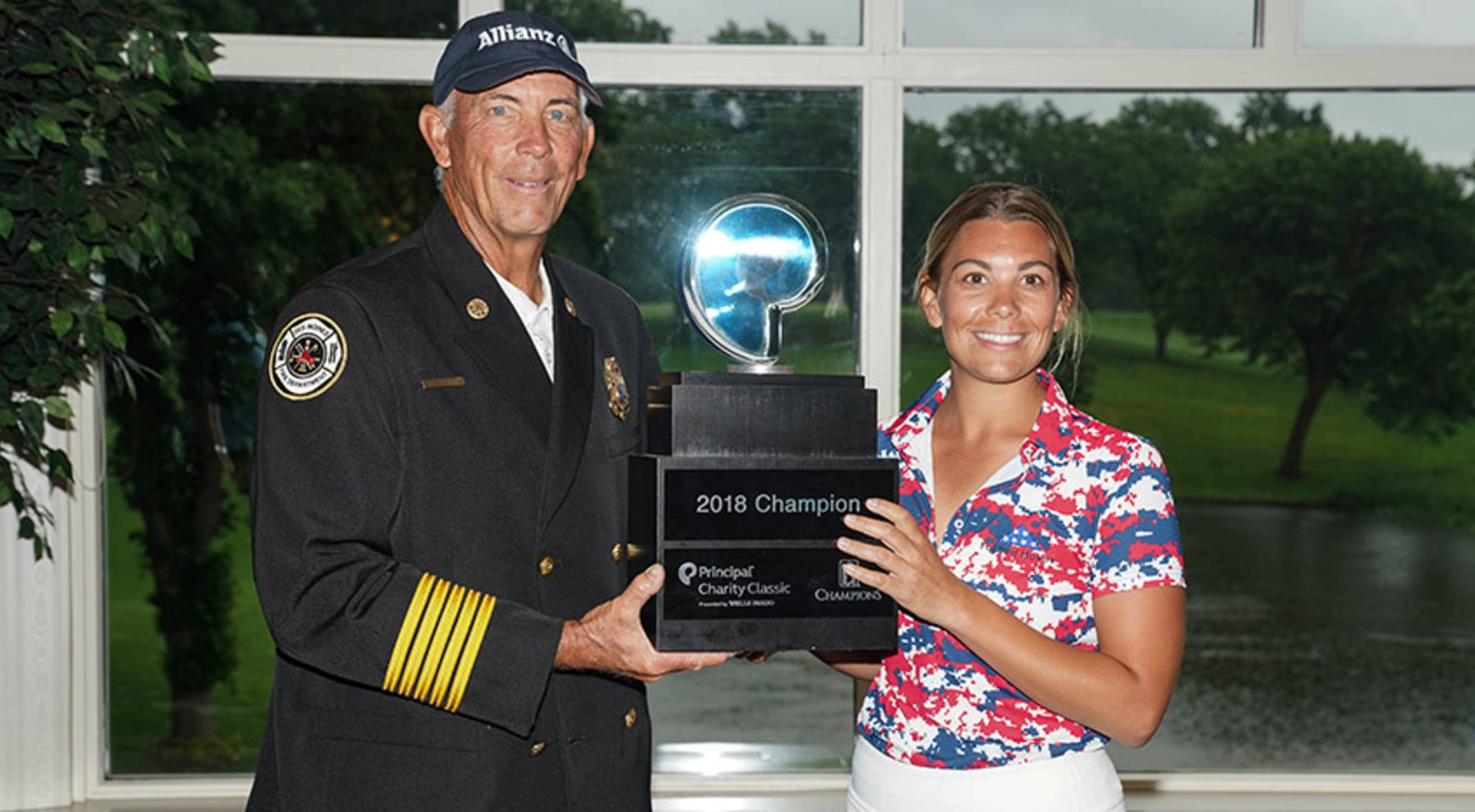 Champion 桌布 壁紙 — 一位微笑的海軍制服男子和一位穿著愛國襯衫的女子在室內共同舉起2018冠軍獎盃，透過他們身後的窗戶可以看到高爾夫球場。
