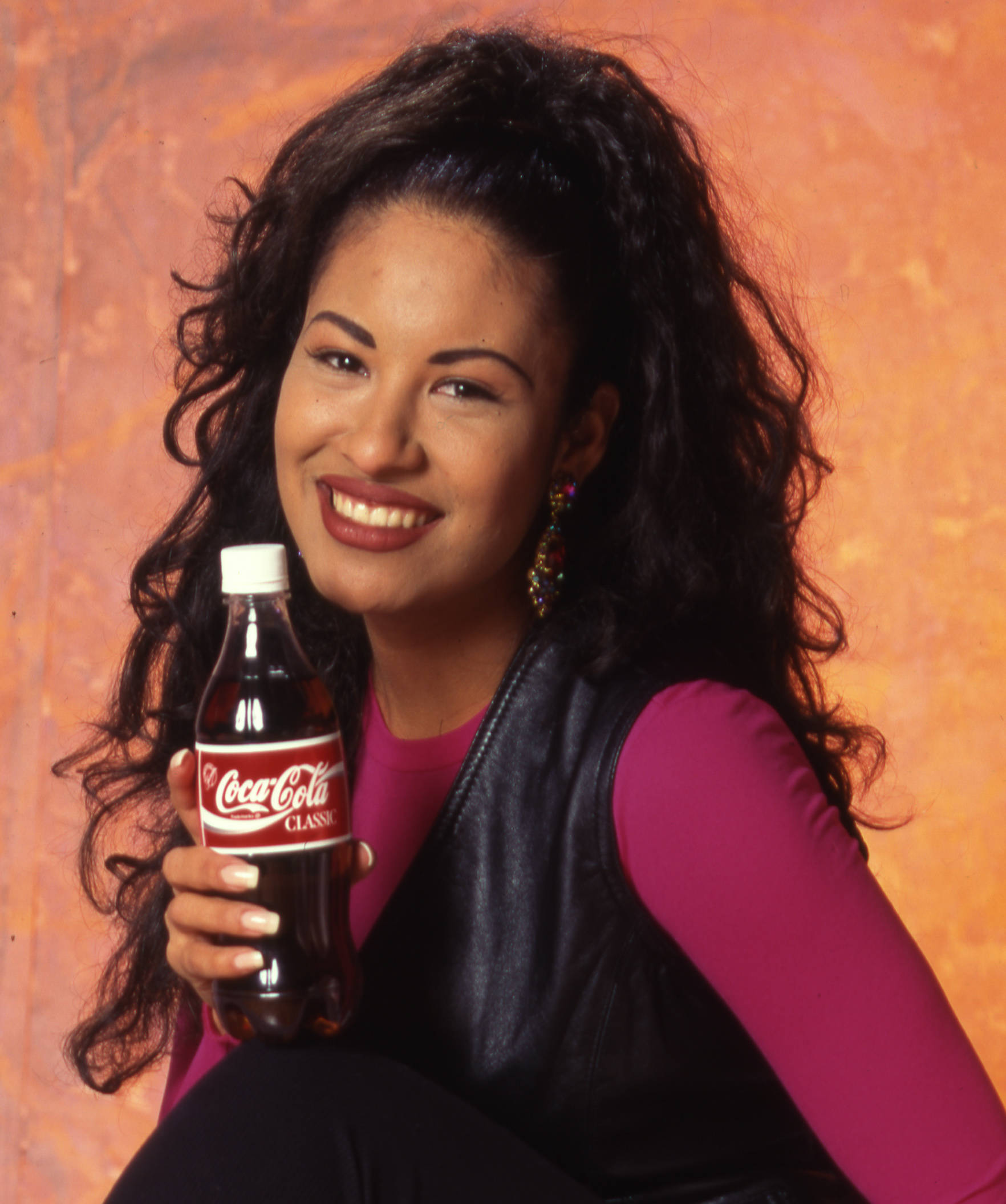 Selena Quintanilla 桌布 壁紙 — 一位微笑的深色捲髮女性，在溫暖的紋理橙色背景前手持可口可樂瓶。