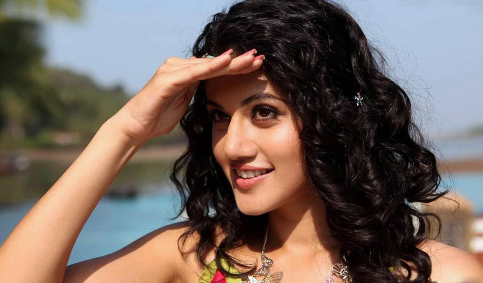 Taapsee Pannu 桌布 壁紙 — 一位微笑的深色捲髮女性在平靜的藍色水體附近遮擋刺眼的陽光。