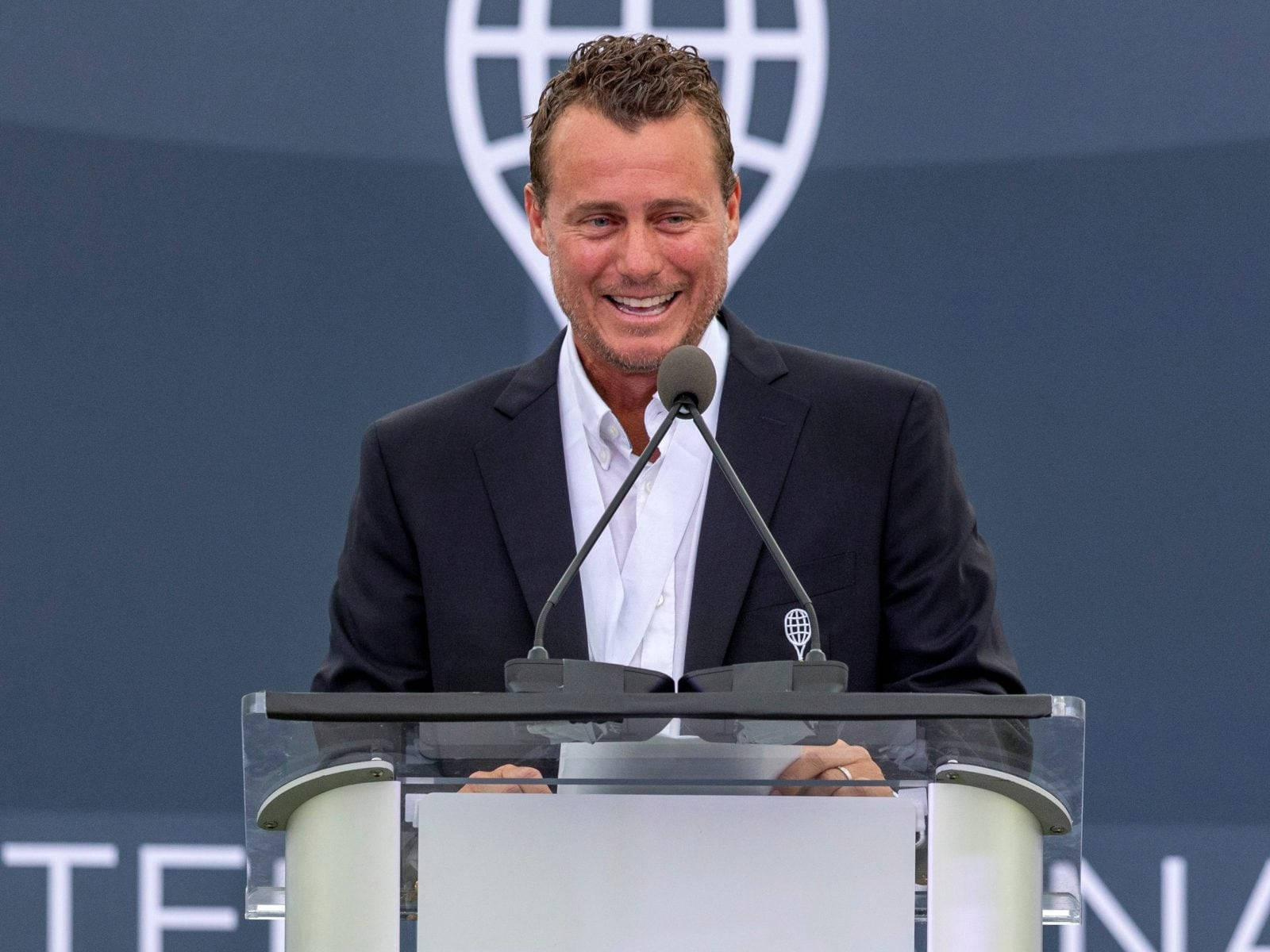 Tennis 桌布 壁紙 — 一位微笑的深色西裝男士站在透明講台後方，講台上有麥克風，背景為藍色並帶有白色地球標誌。