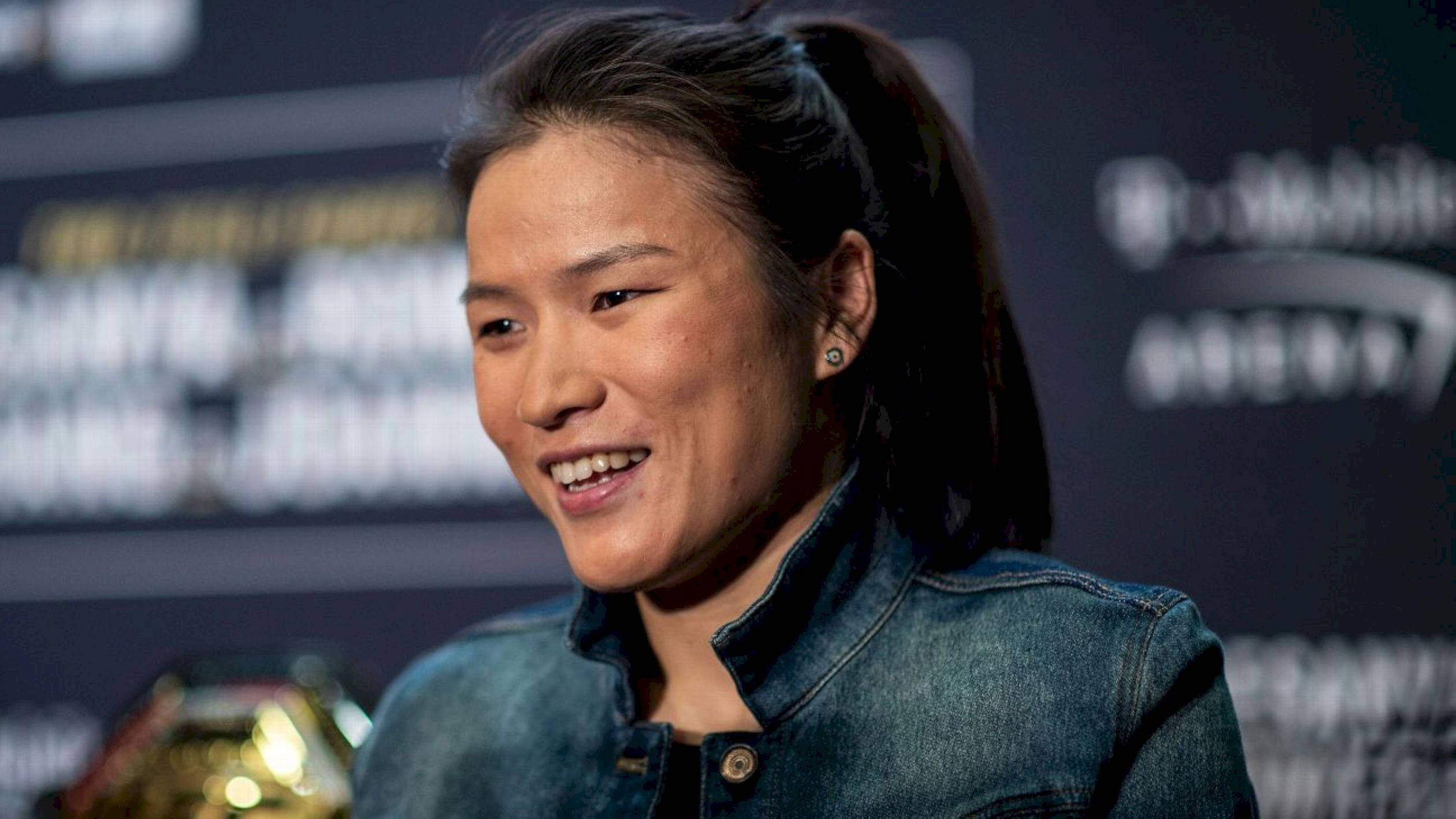 Athletic 桌布 壁紙 — 一位微笑的深色頭髮馬尾辮女性，在背後有模糊標誌的新聞活動中穿著牛仔外套。