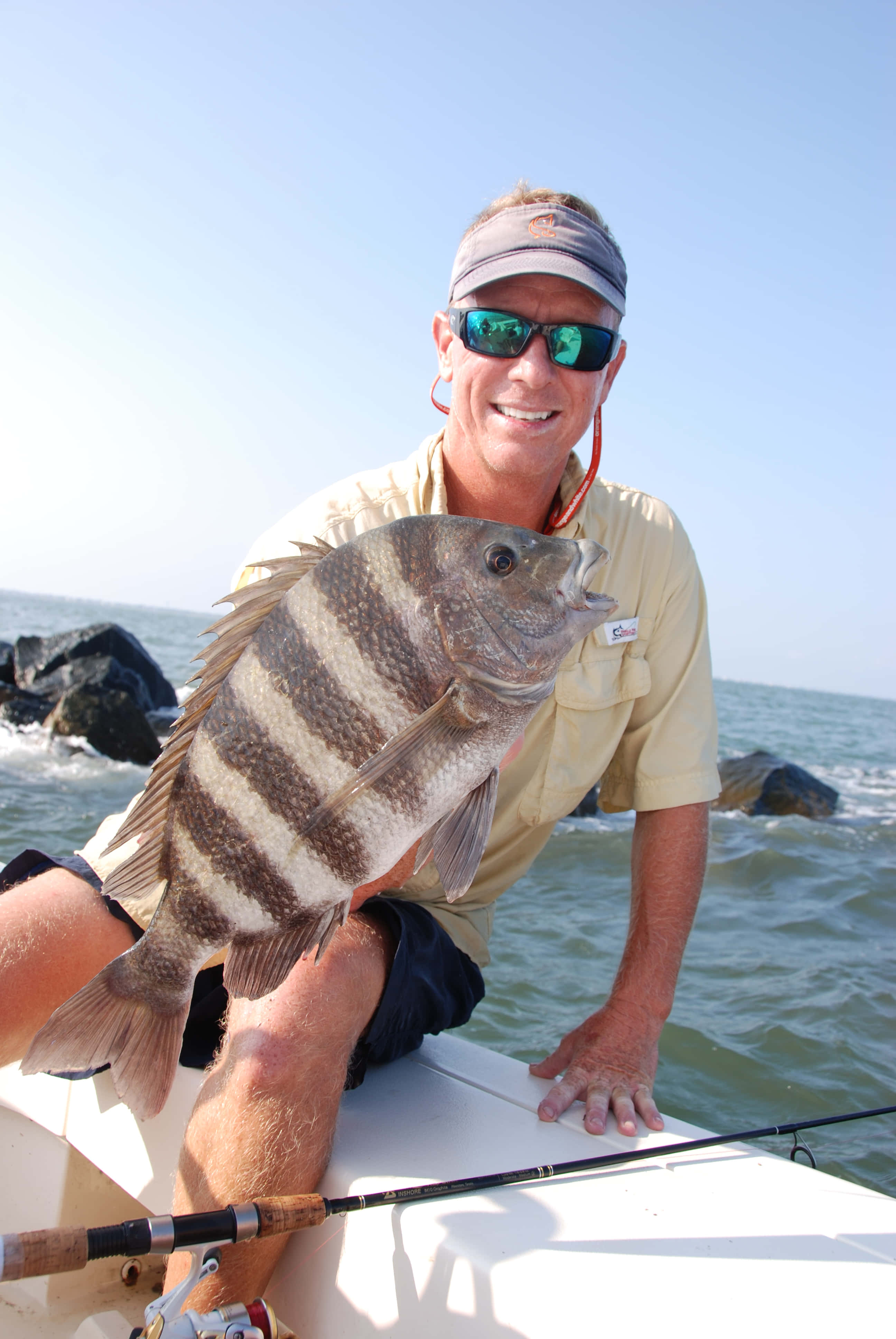 Sheepshead Fish 桌布 壁紙 — 一位微笑的漁夫在船上自豪地拿著一條大條紋魚，背景是海洋和岩石，在明亮的陽光下。