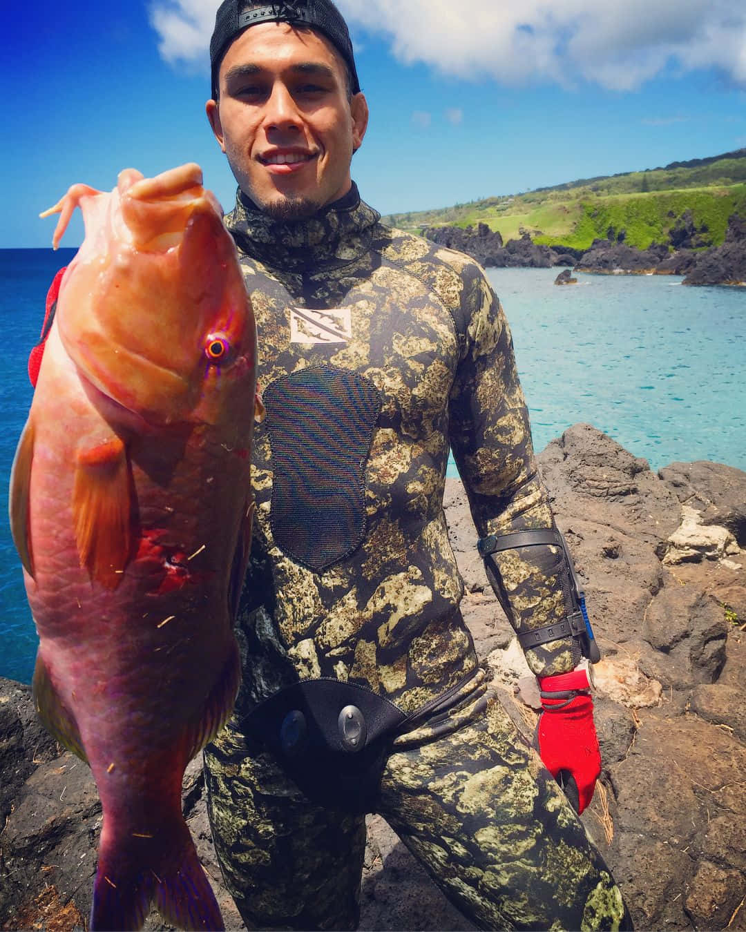 釣魚 桌布 壁紙 — 一位微笑的漁夫穿著迷彩潛水衣，在陽光明媚的岩石海岸上自豪地拿著一條大粉紅魚，背景是碧綠的海水和綠色的山丘。