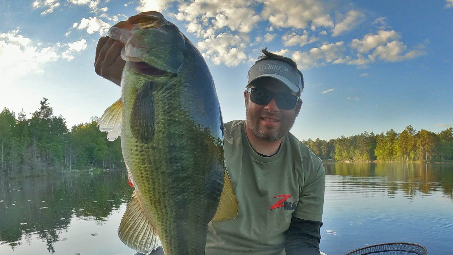 Largemouth Bass 桌布 壁紙 — 一位微笑的漁夫自豪地抱著一條巨大的鱸魚，背景是寧靜的湖泊，有樹木和部分多雲的天空。