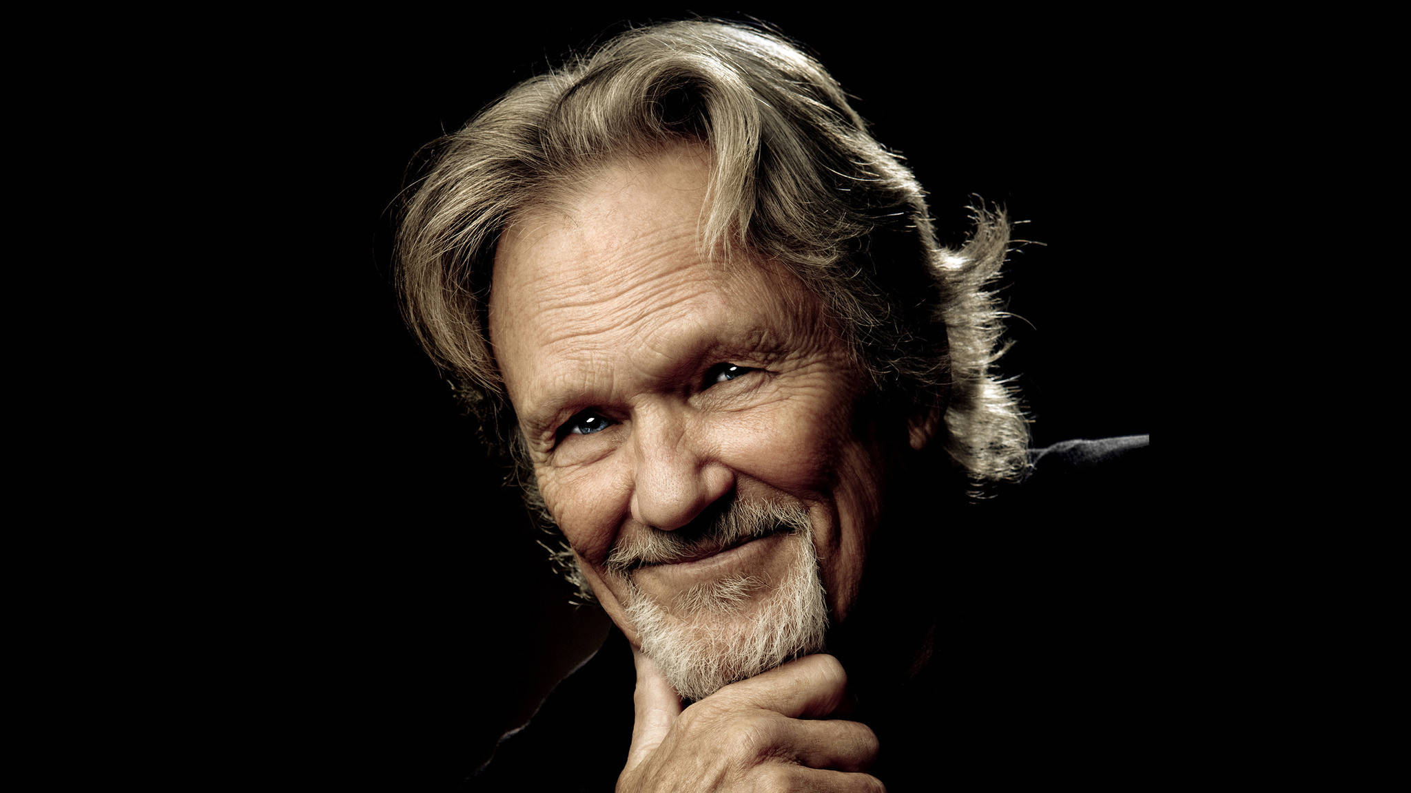 Kris Kristofferson 桌布 壁紙 — 一位微笑的灰白頭髮和鬍鬚的年長男士的頭像特寫，背景為黑色。