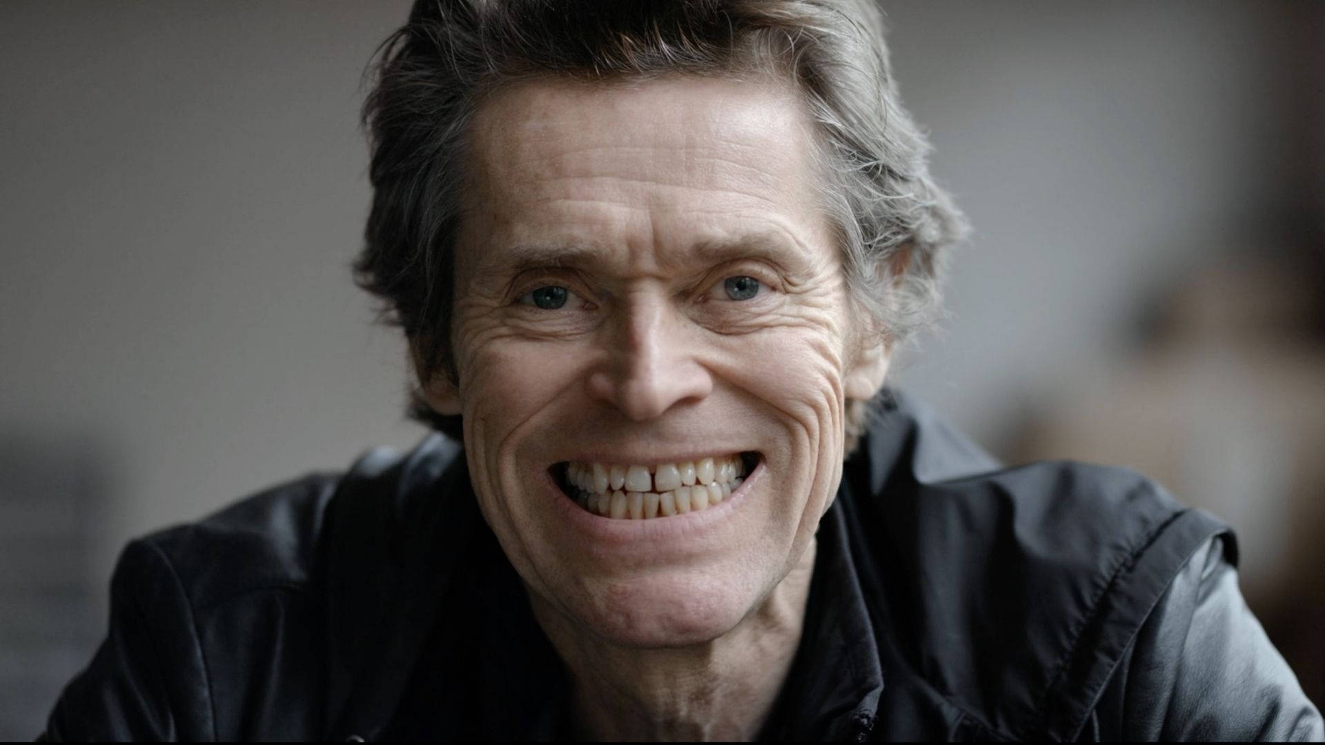 Willem Dafoe 桌布 壁紙 — 一位微笑的灰髮藍眼老年男子穿著黑色外套的特寫肖像。