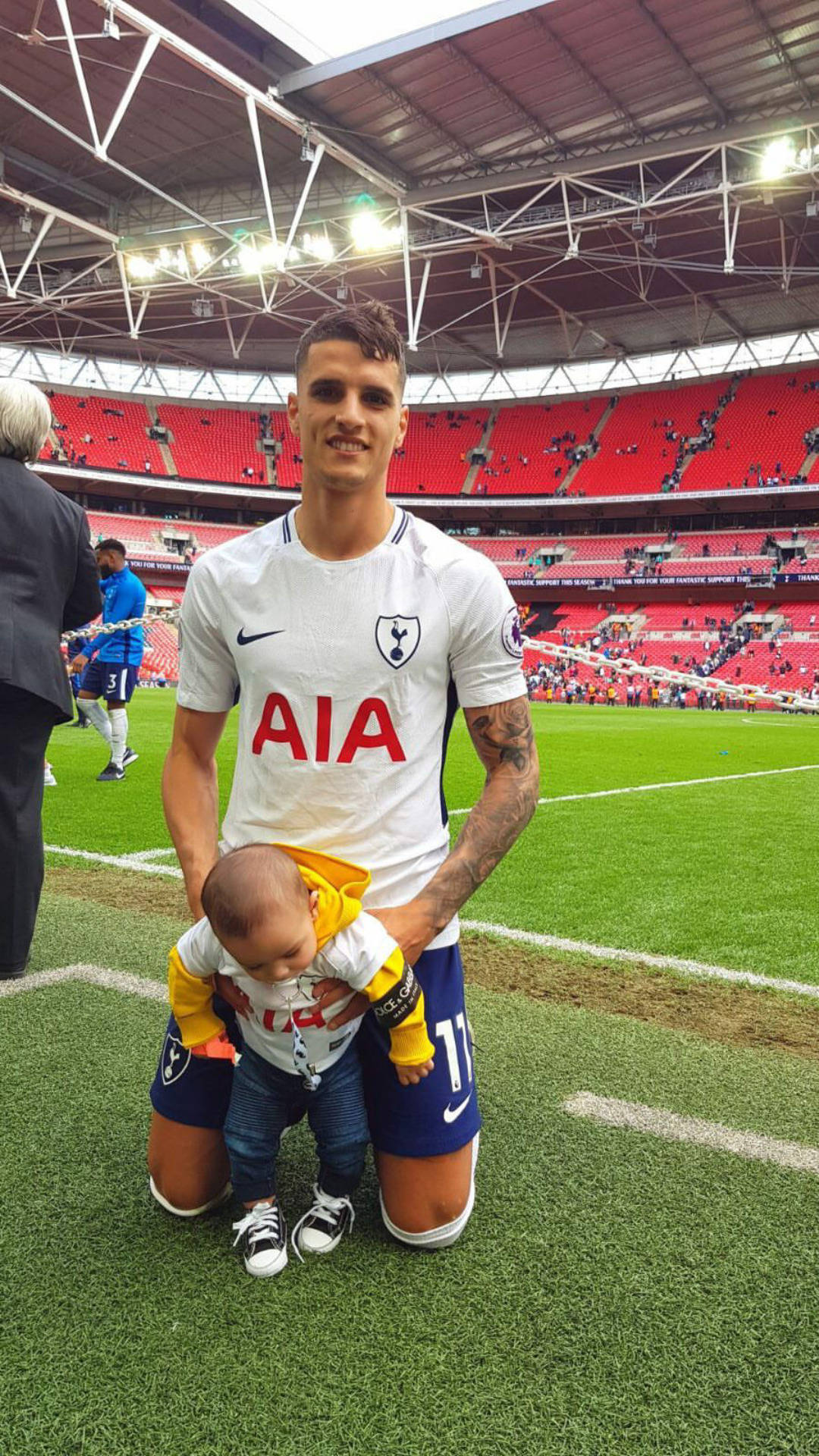 erik lamela 桌布 壁紙 — 一位微笑的熱刺足球員在紅色座位的球場上跪下，抱著一個嬰兒。