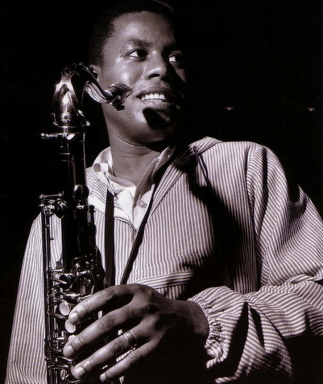 Wayne Shorter 桌布 壁紙 — 一位微笑的爵士薩克斯風手穿著條紋襯衫，在戲劇性的舞台燈光下拿著他的樂器。