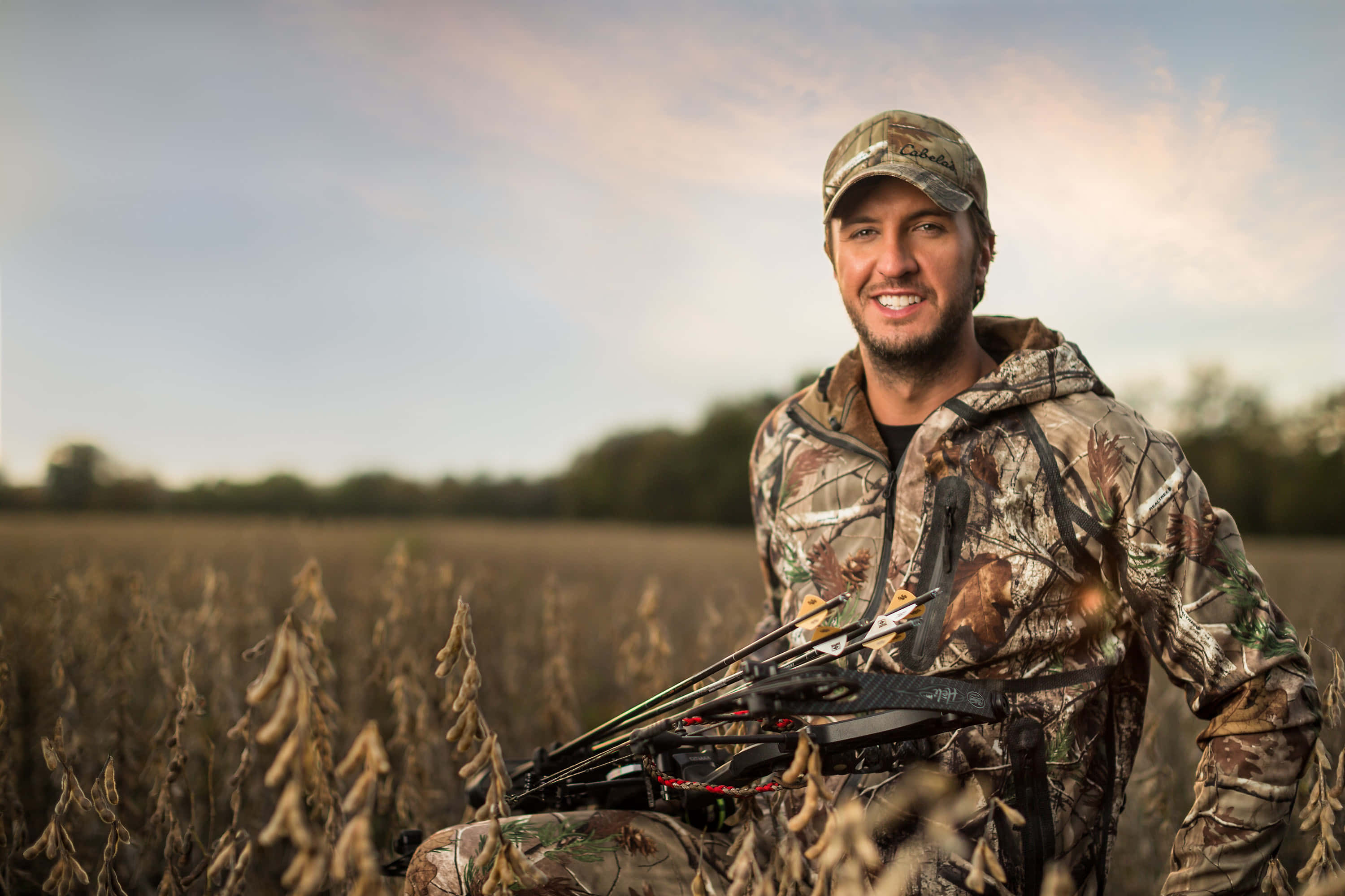 Luke Bryan 桌布 壁紙 — 一位微笑的獵人身穿迷彩裝備，在夕陽下站在乾燥的大豆田中手持弓箭。