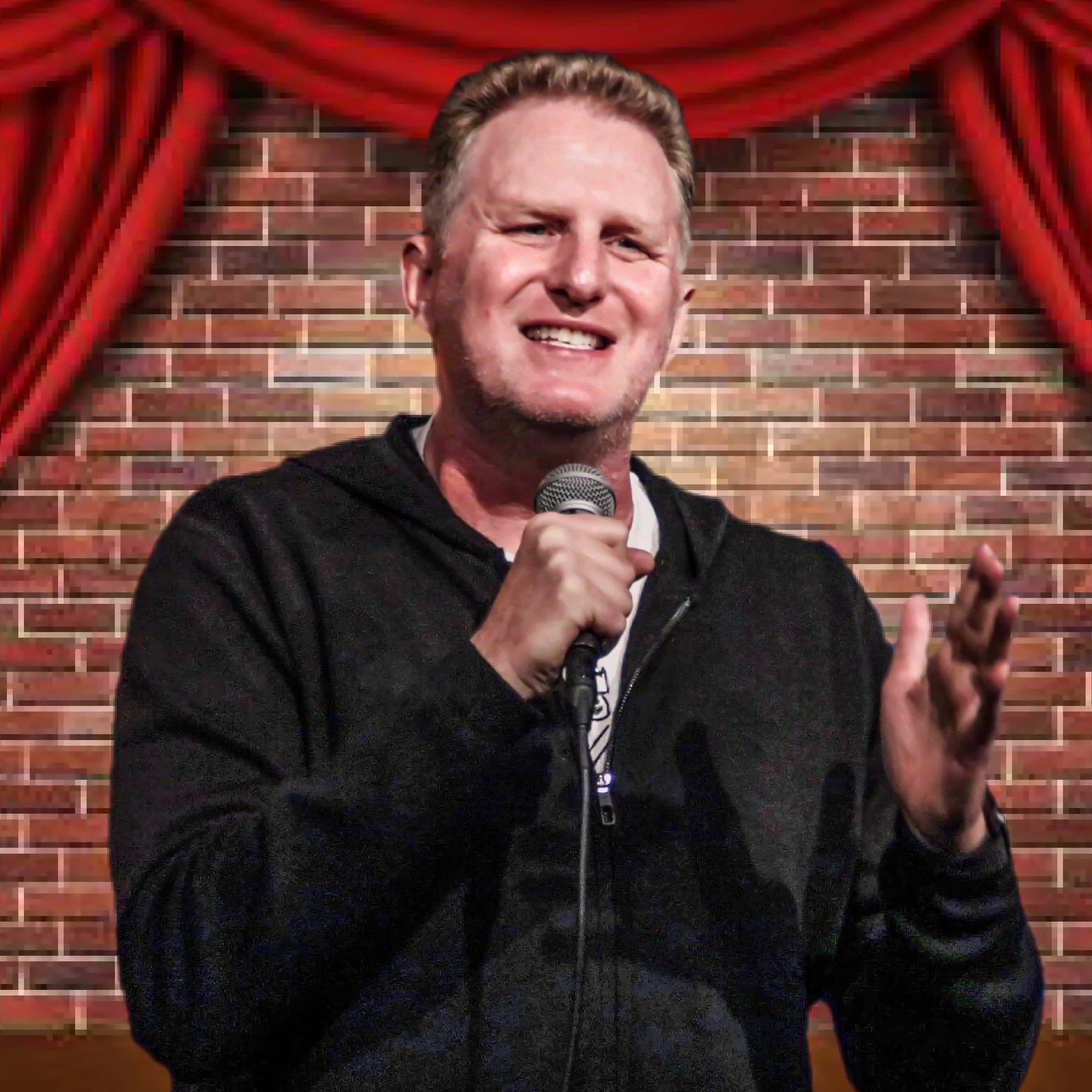 Michael Rapaport 桌布 壁紙 — 一位微笑的男喜劇演員在舞台上拿著麥克風，身後有紅色窗簾和磚牆。