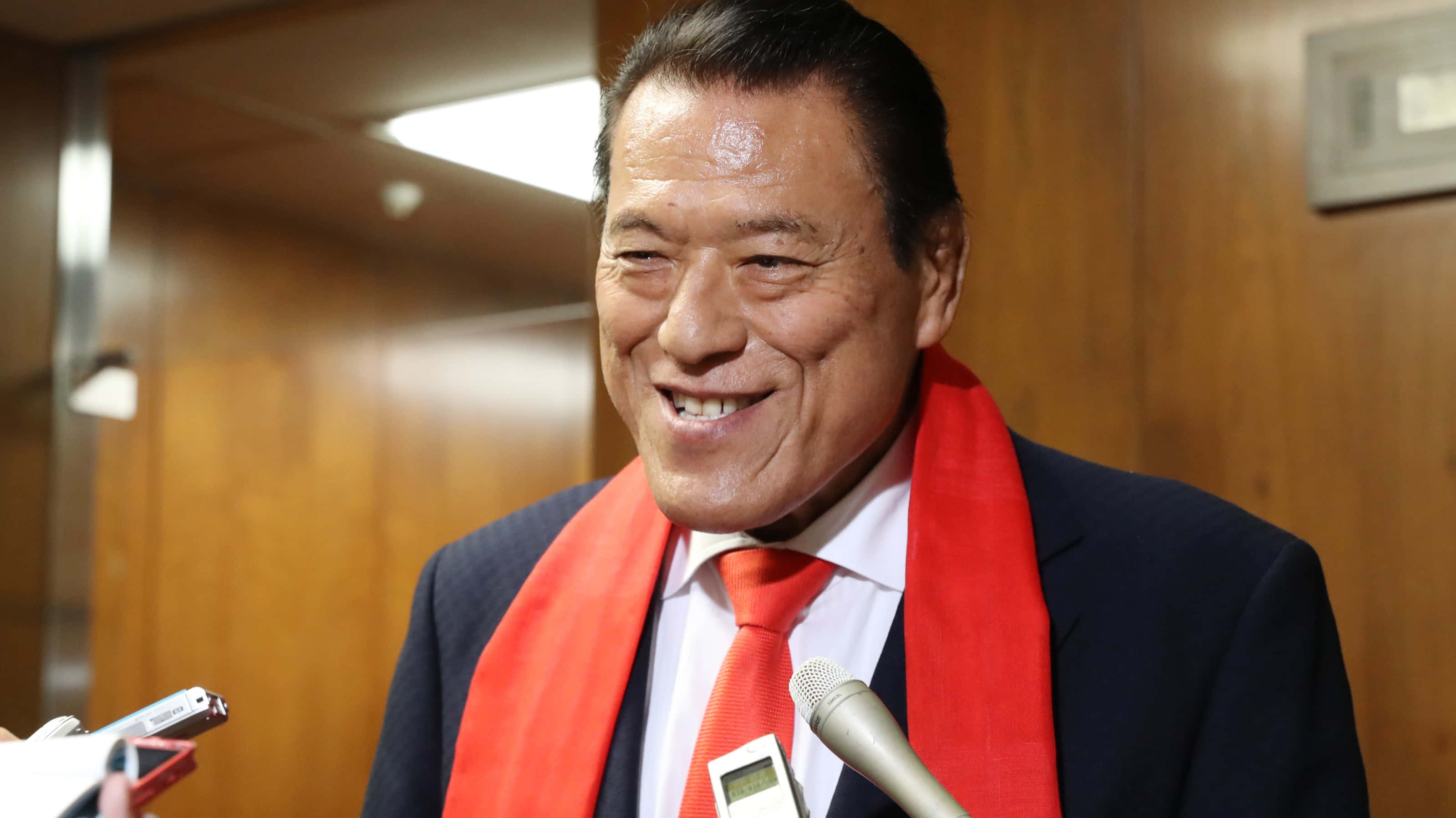 摔角 桌布 壁紙 — 一位微笑的男士，身穿西裝打紅色領帶，站在木牆板房間的麥克風前面。