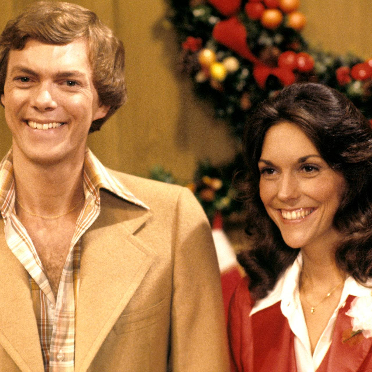 Carpenters 桌布 壁紙 — 一位微笑的男士和女士穿著1970年代的服裝，在節日聖誕花環前溫馨地擺姿勢。