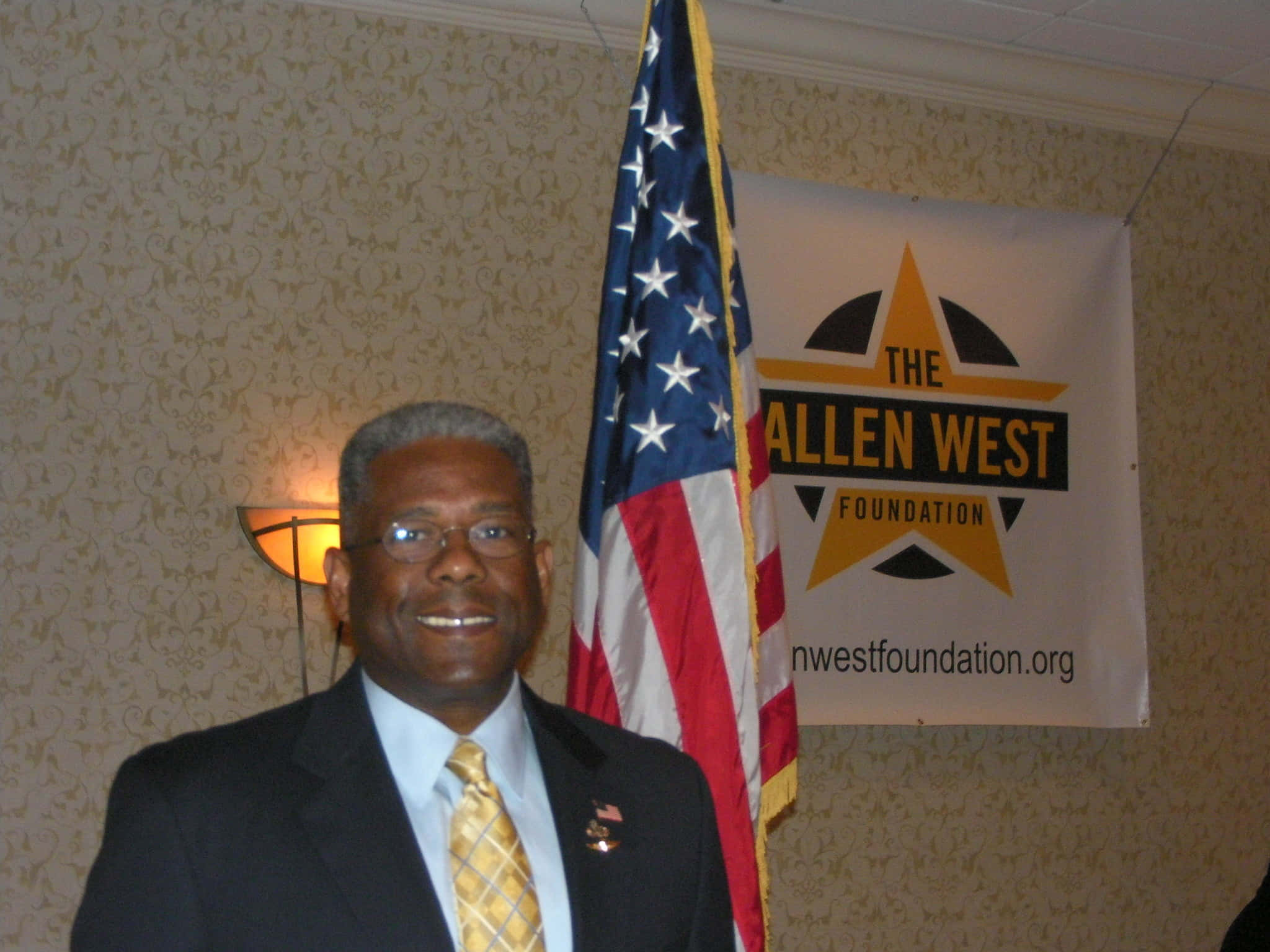 Famous 桌布 壁紙 — 一位微笑的男士穿著西裝打領帶，站在美國國旗和The Allen West Foundation的橫幅前面。