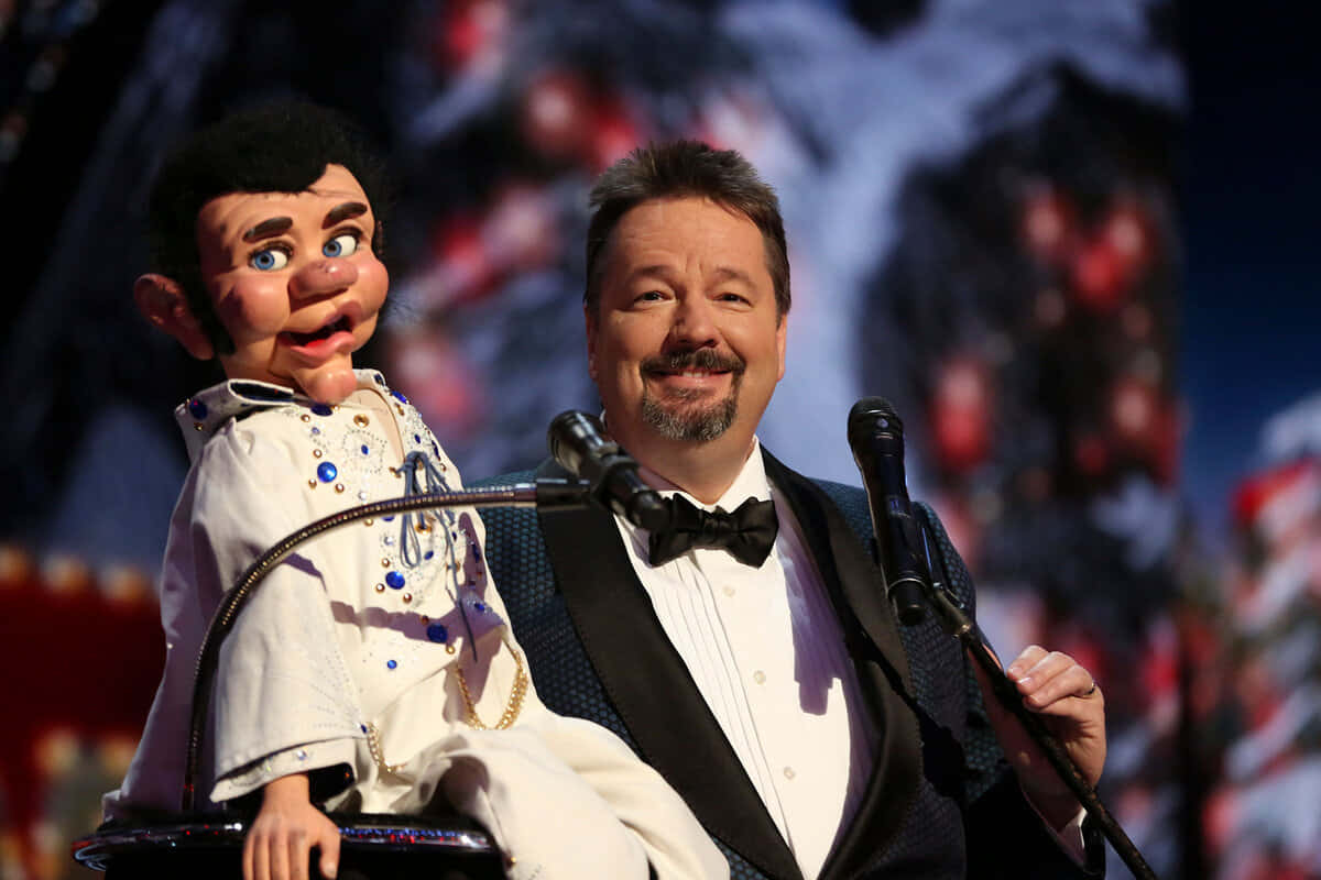puppet 桌布 壁紙 — 一位微笑的男士身穿燕尾服，在舞台上與身穿閃亮白色連身衣的貓王普雷斯利人偶一起表演。